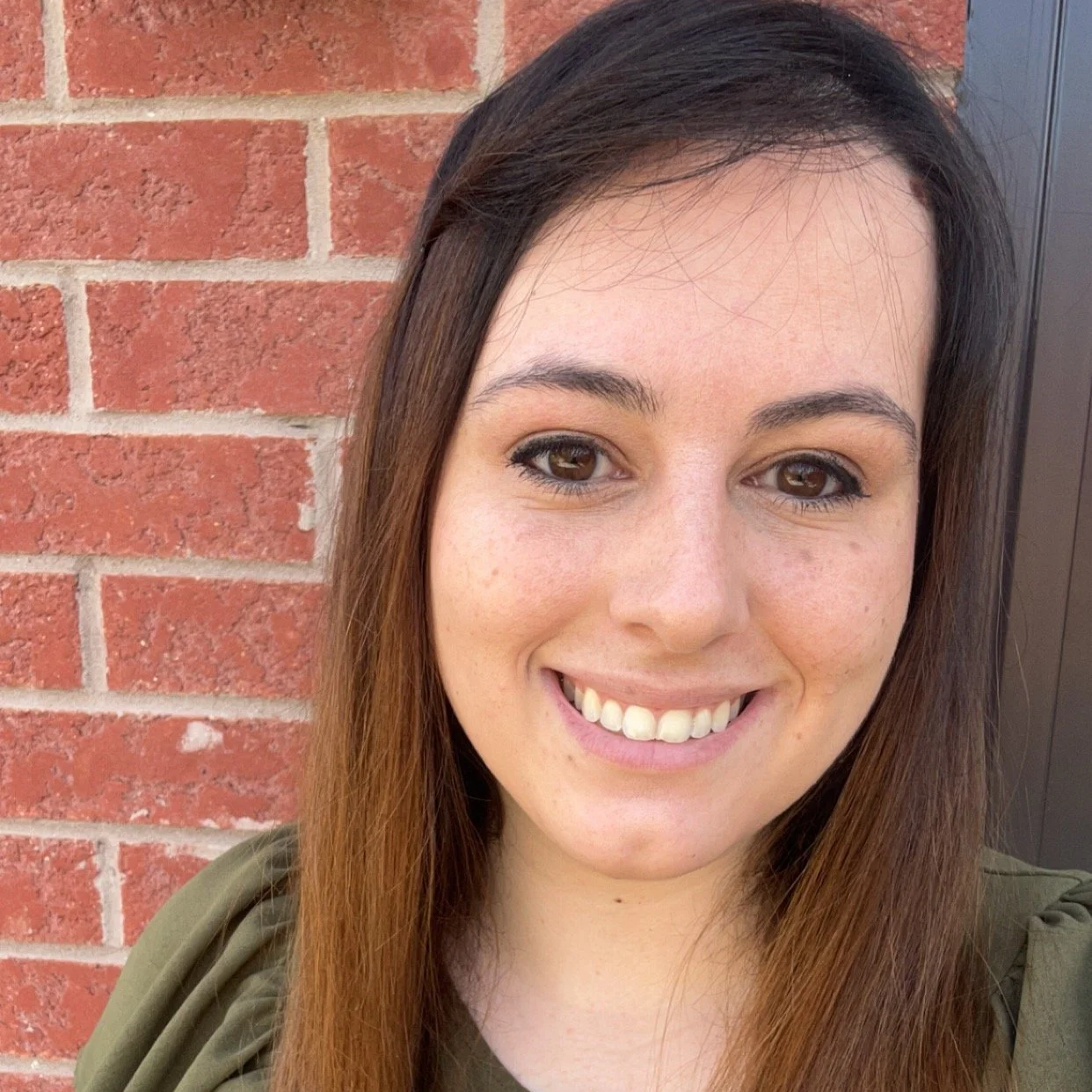 Close-up of a smiling woman with long brown hair, brown eyes, and freckles, standing in front of a red brick wall.