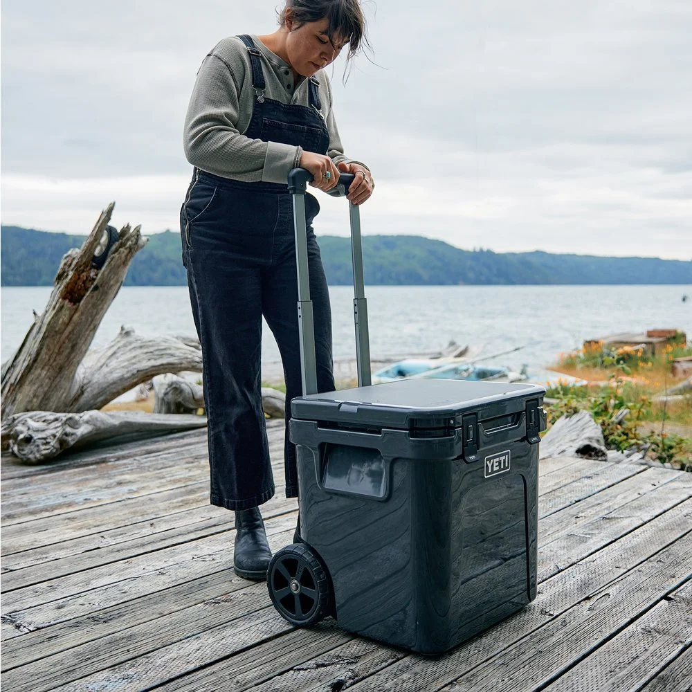 Woman standing on a wooden dock next to a lake, extending a handle on a black YETI cooler with wheels.