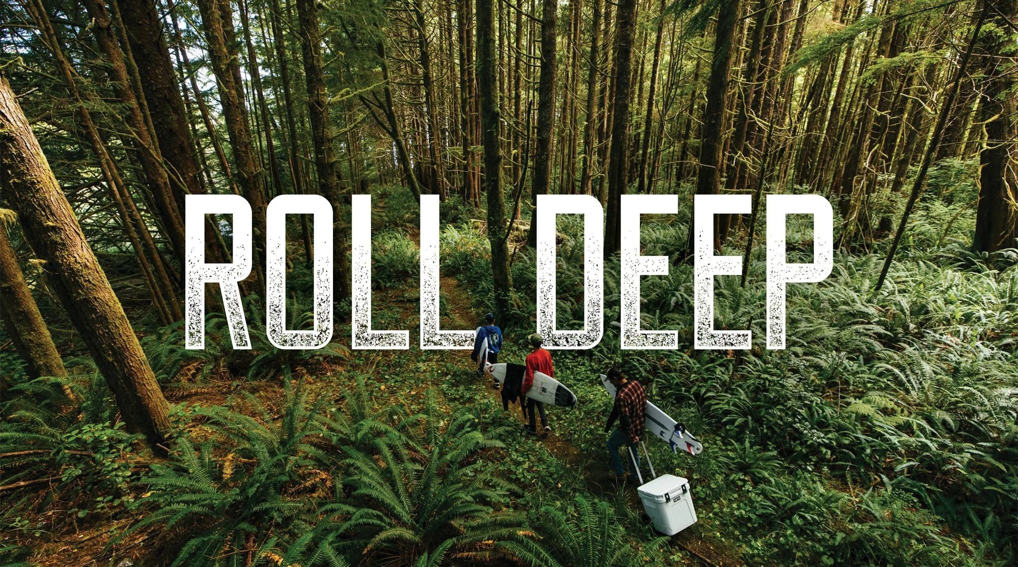 A group of four people with surfboards and rolling a YETI wheeled cooler walking through a lush, green forest with tall trees and dense foliage, with the words 'ROLL DEEP' overlayed in white distressed text.