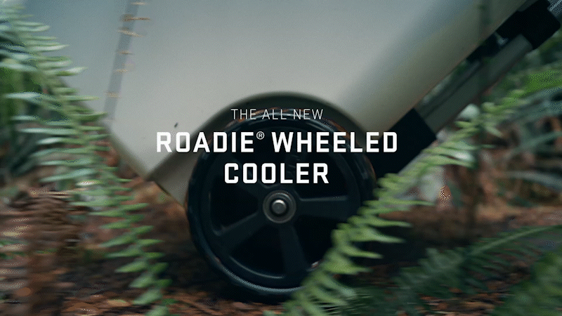 Close-up of a wheel on a cooler rolling along a dirt path with text indicating it is a Roadie Wheeled Cooler, surrounded by soil and green plants.