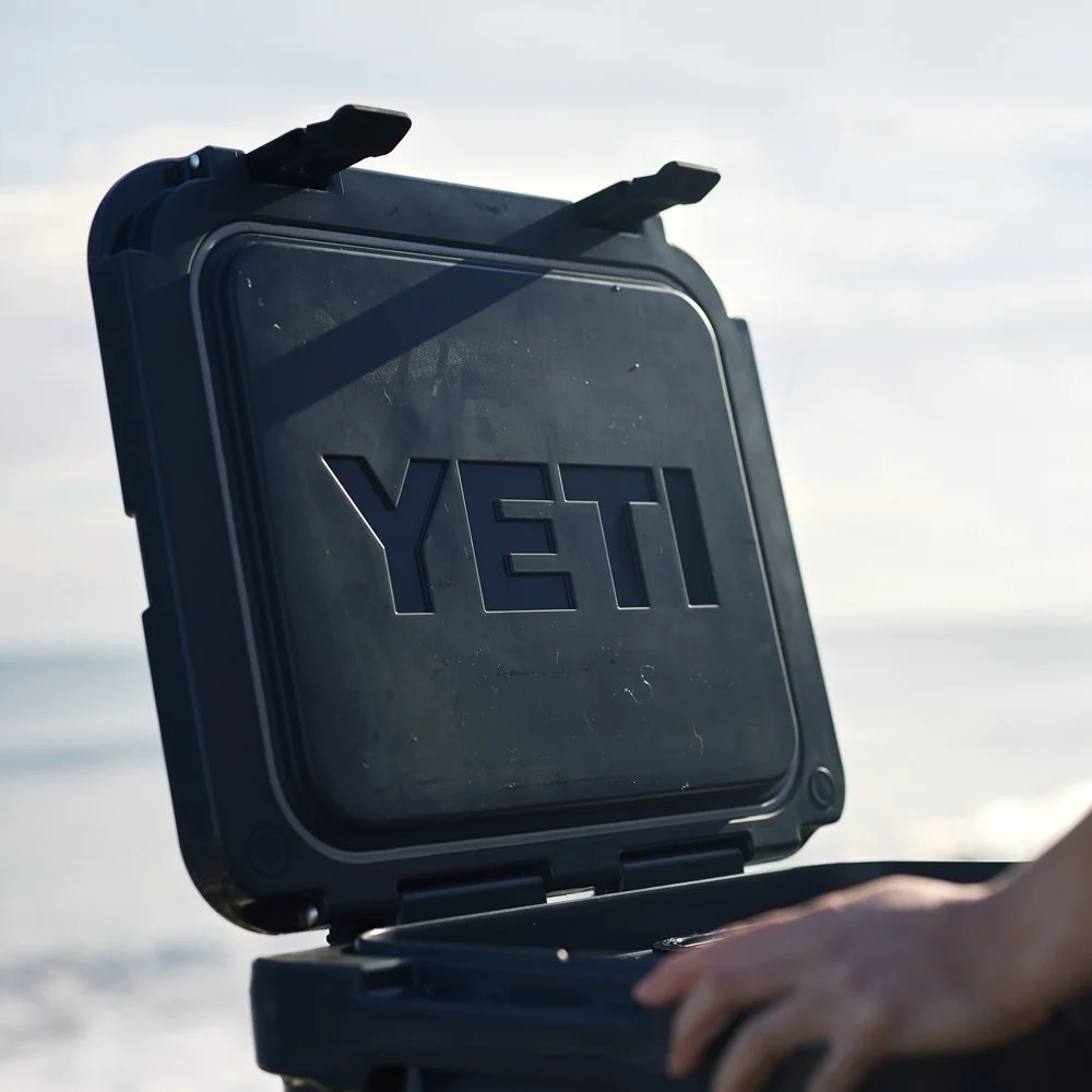 A black YETI cooler with its lid open near a beach, with a person's hand resting on the cooler's edge.