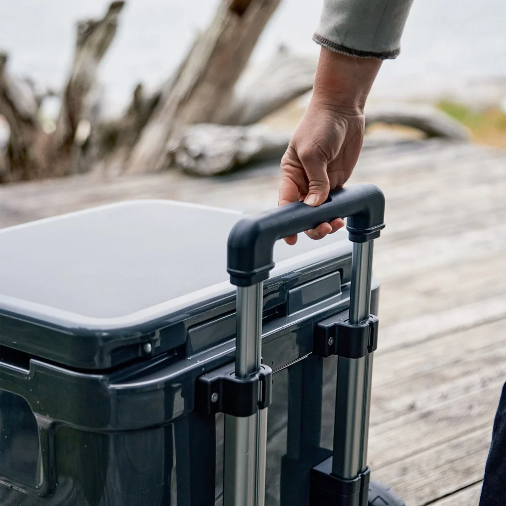 A hand gripping the handle of a black wheelie cart on a wooden dock, with a blurred background of driftwood and water.