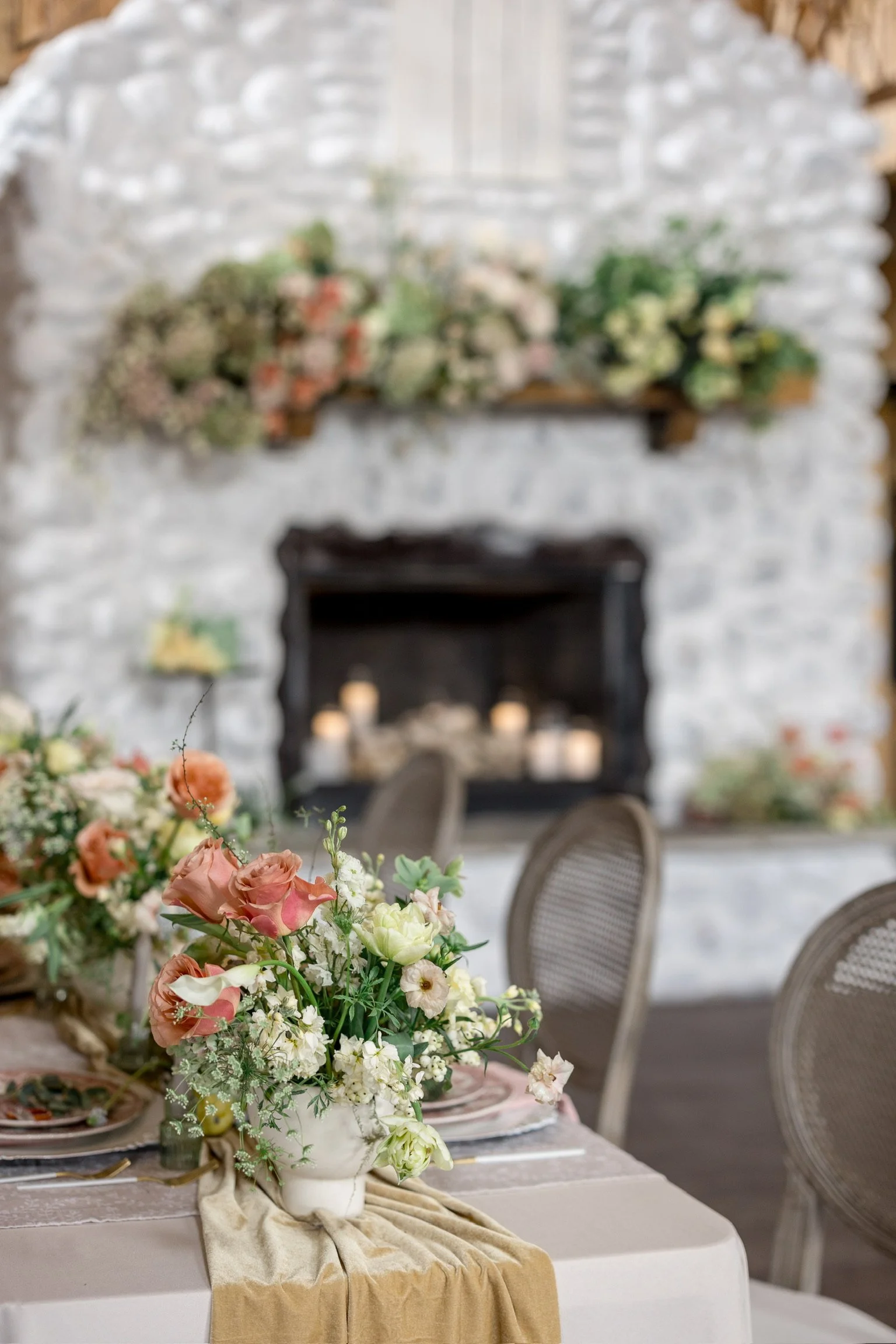 A wedding reception table decorated with a large floral arrangement of pink, white, and green flowers, beneath a champagne-colored tablecloth, with blurred chairs around and a fireplace with candles and floral decor in the background.