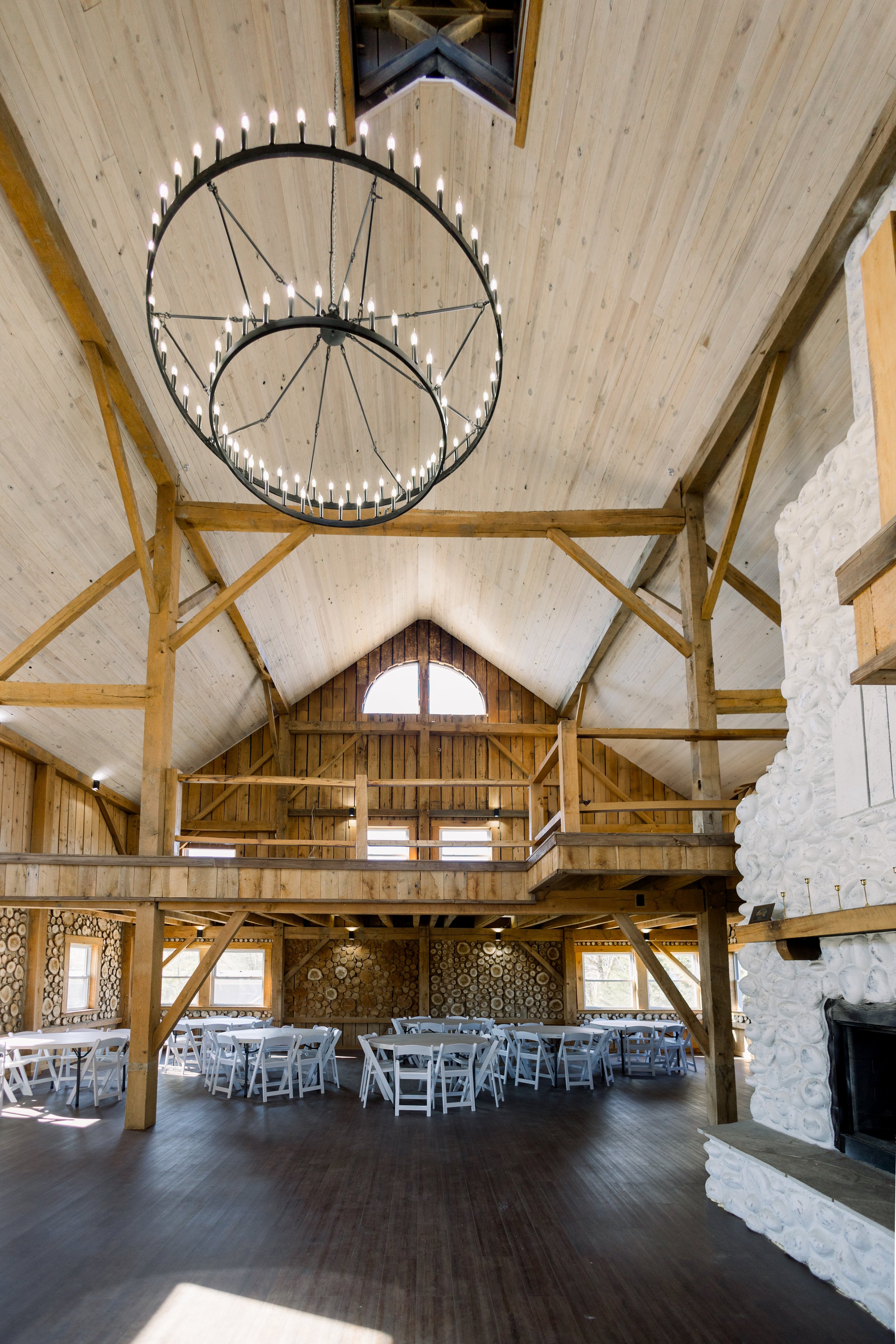 Interior of a rustic event hall with wooden beams, high vaulted ceiling, white tables and chairs, a large hanging chandelier, stone fireplace, and natural light coming through windows.
