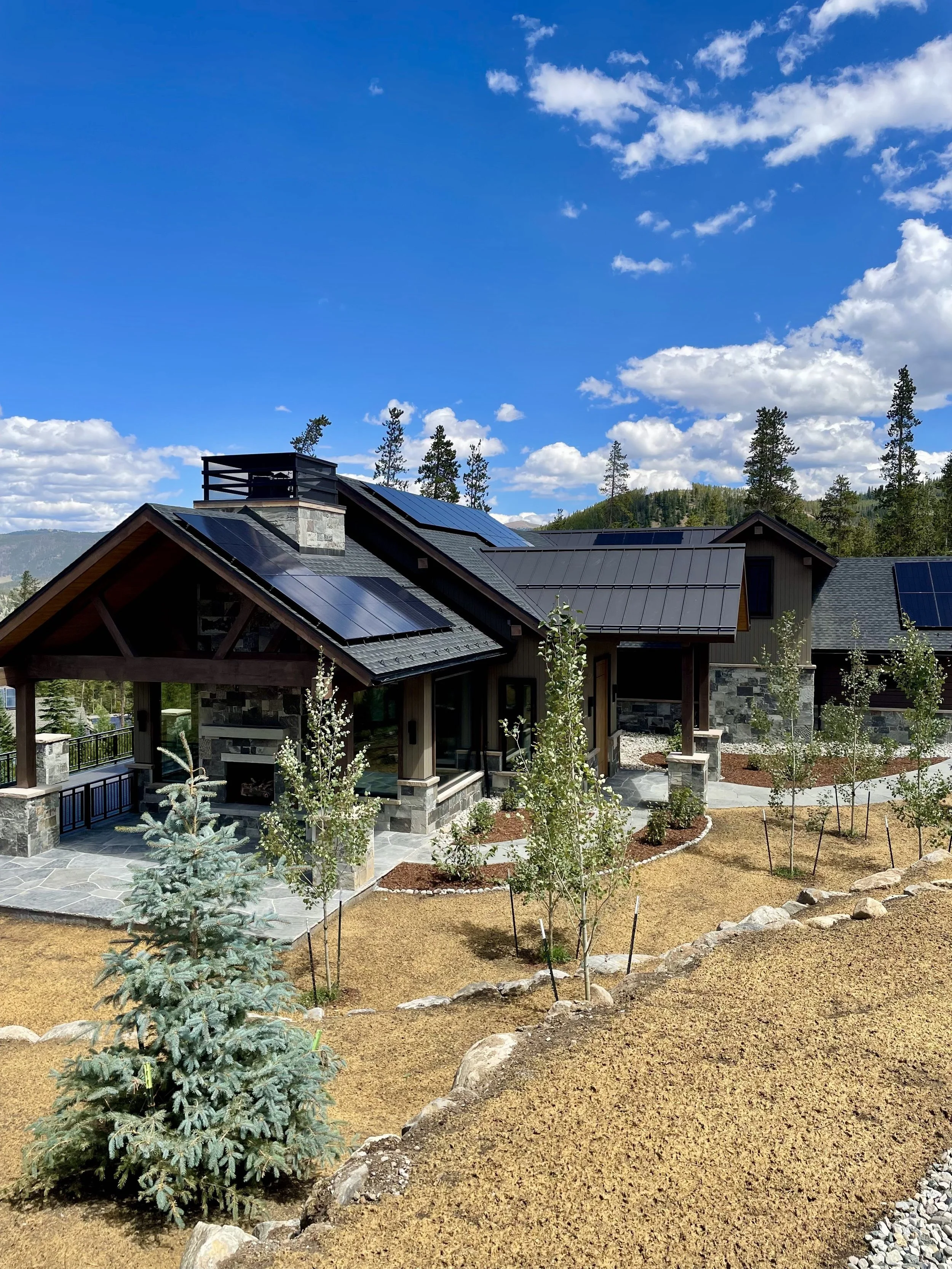 A house with solar panels on the roof, surrounded by a landscaped yard with young trees, rocks, and a mountain backdrop under a partly cloudy sky.