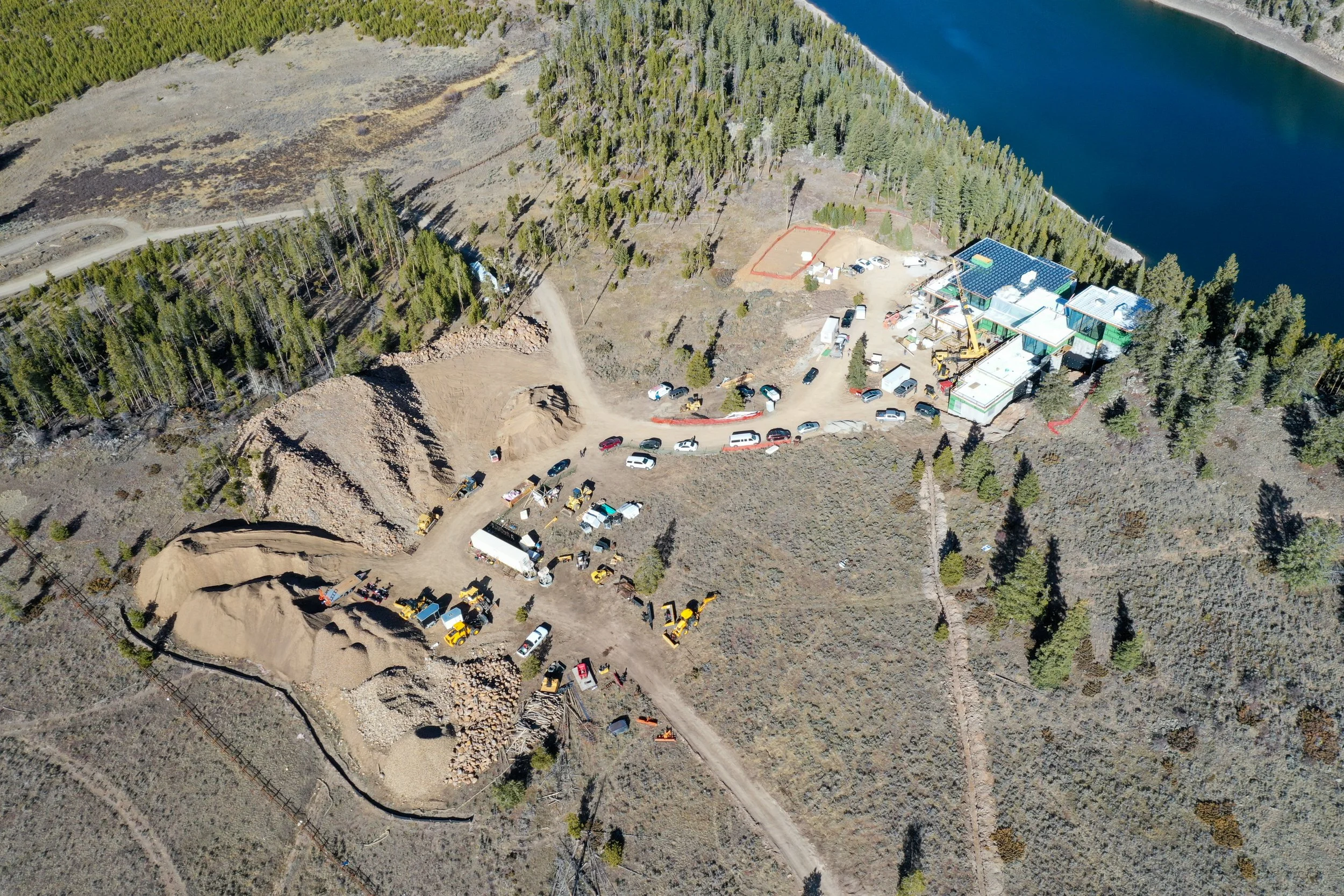 Aerial view of a construction site near a body of water with cleared land, construction equipment, and parked vehicles amid a forested area.