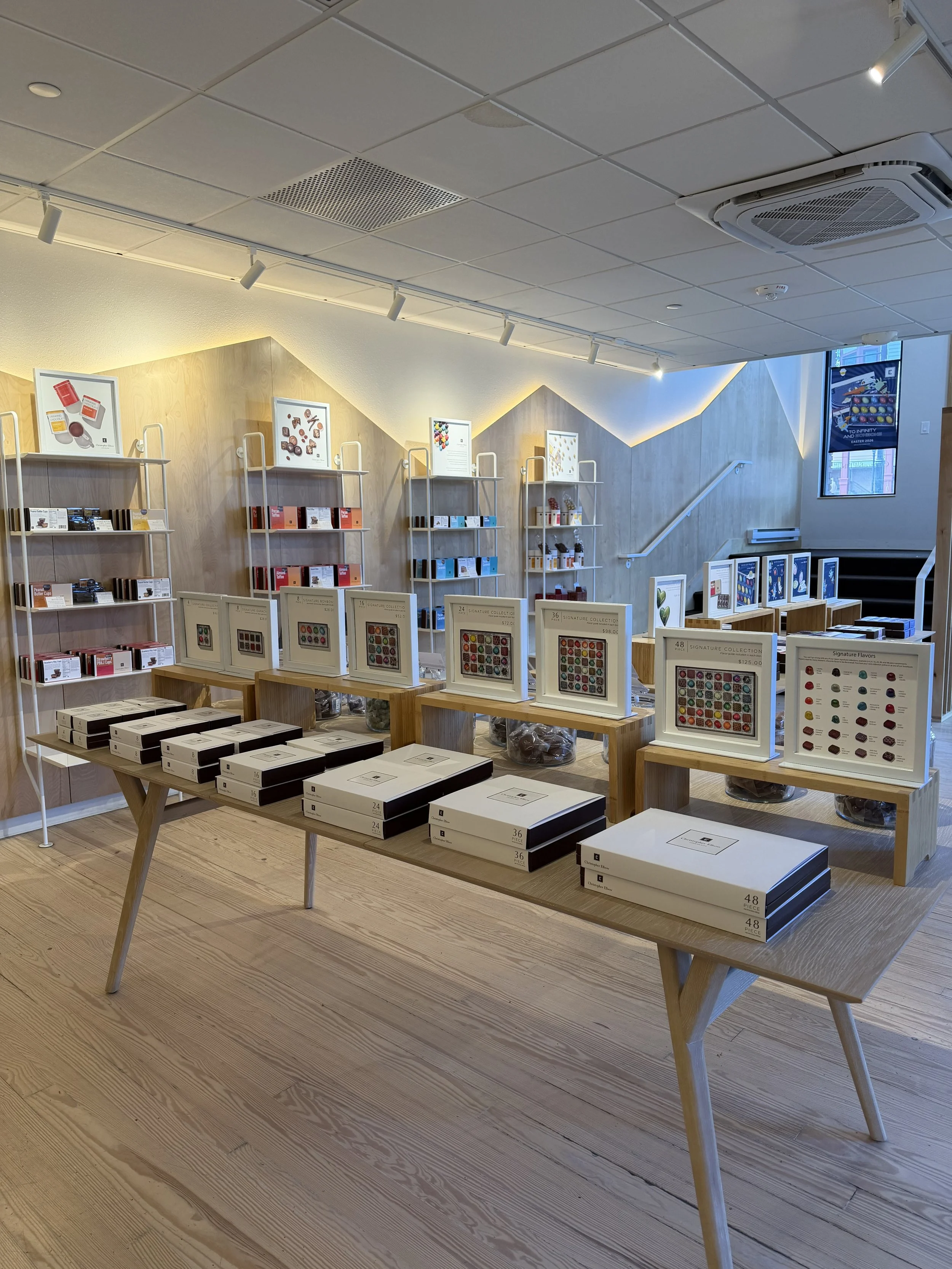Interior of a store or gallery displaying jewelry or accessories, with tables and shelves holding boxed items, framed charts showing different product options, and a staircase leading to an upper level.