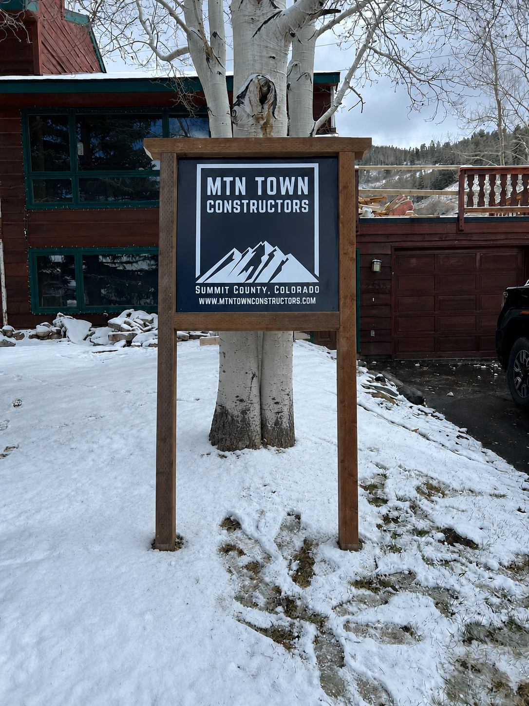 A sign for MTN TOWN CONSTRUCTORS in Summit County, Colorado, standing in snow in front of a tree and a wooden house with red siding and green window frames.