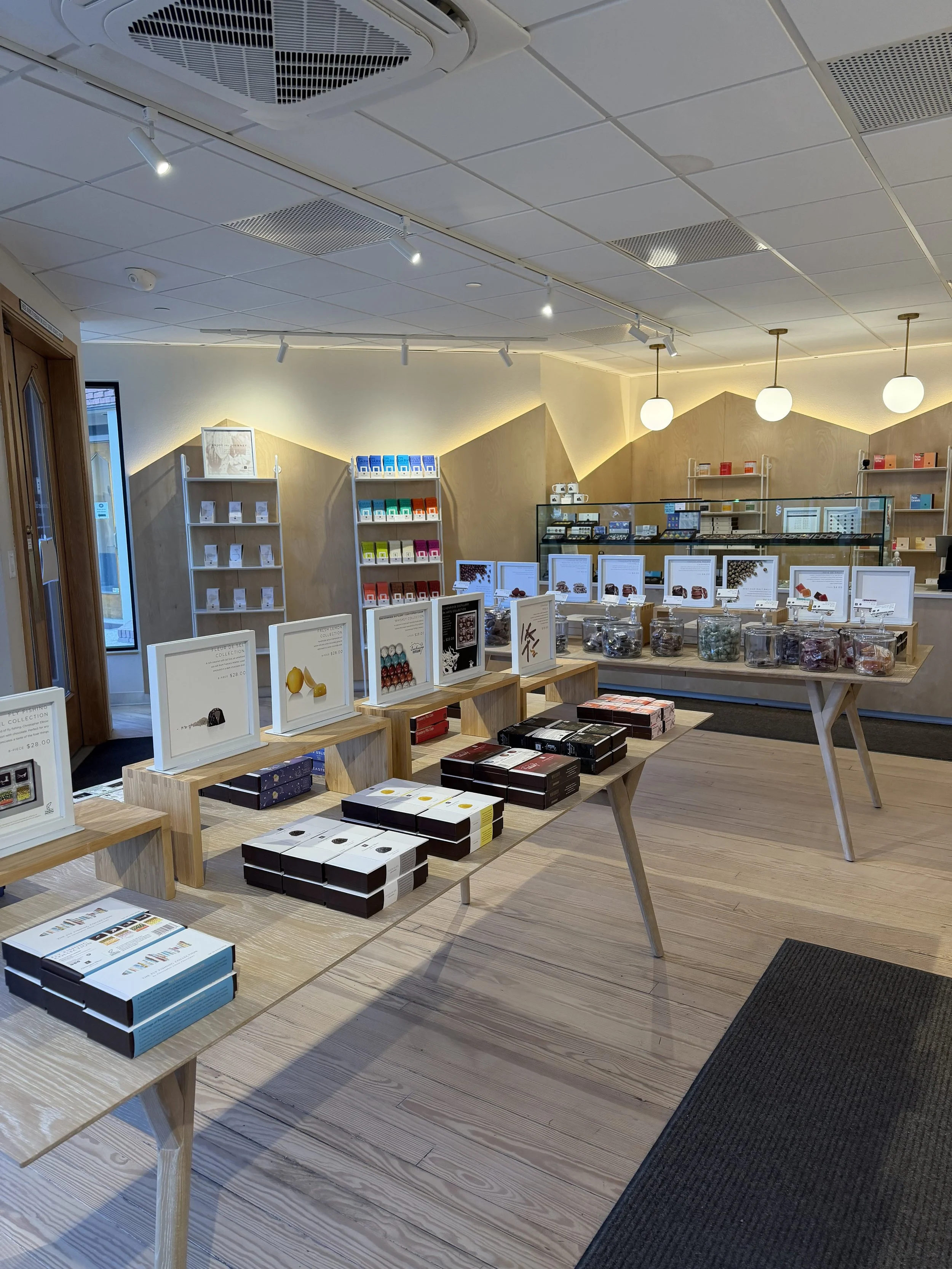 Interior of a retail store with display tables, shelves of colorful boxes of chocolates on light wood flooring and ceiling lights.