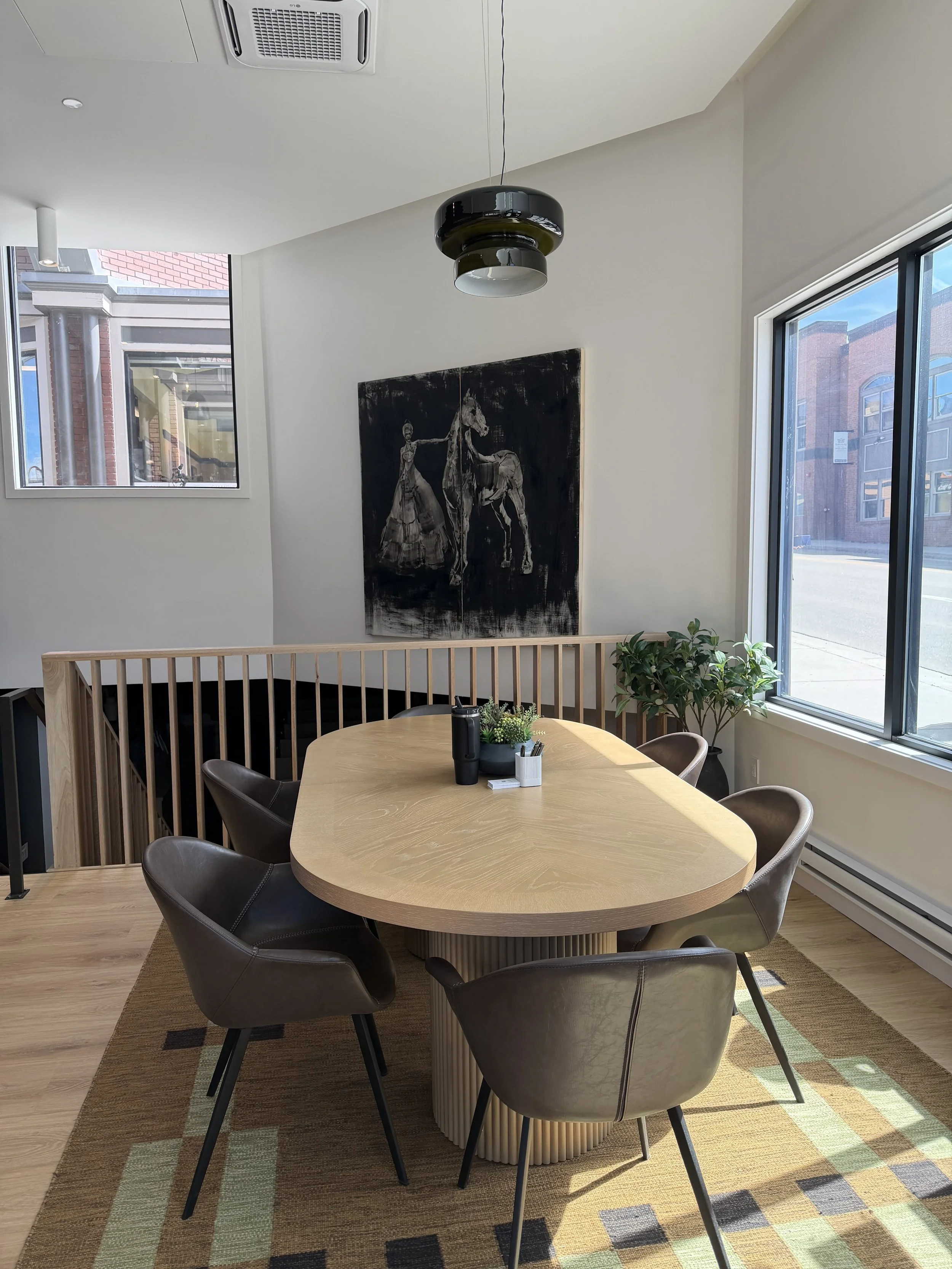 A modern light wood oval table surrounded by six tan leather chairs. There is a small potted plant and a black water bottle on the table. The room has large windows allowing natural light with views of city buildings, and a black and white painting of a woman and a horse on the wall. A ceiling light fixture hangs from the ceiling.