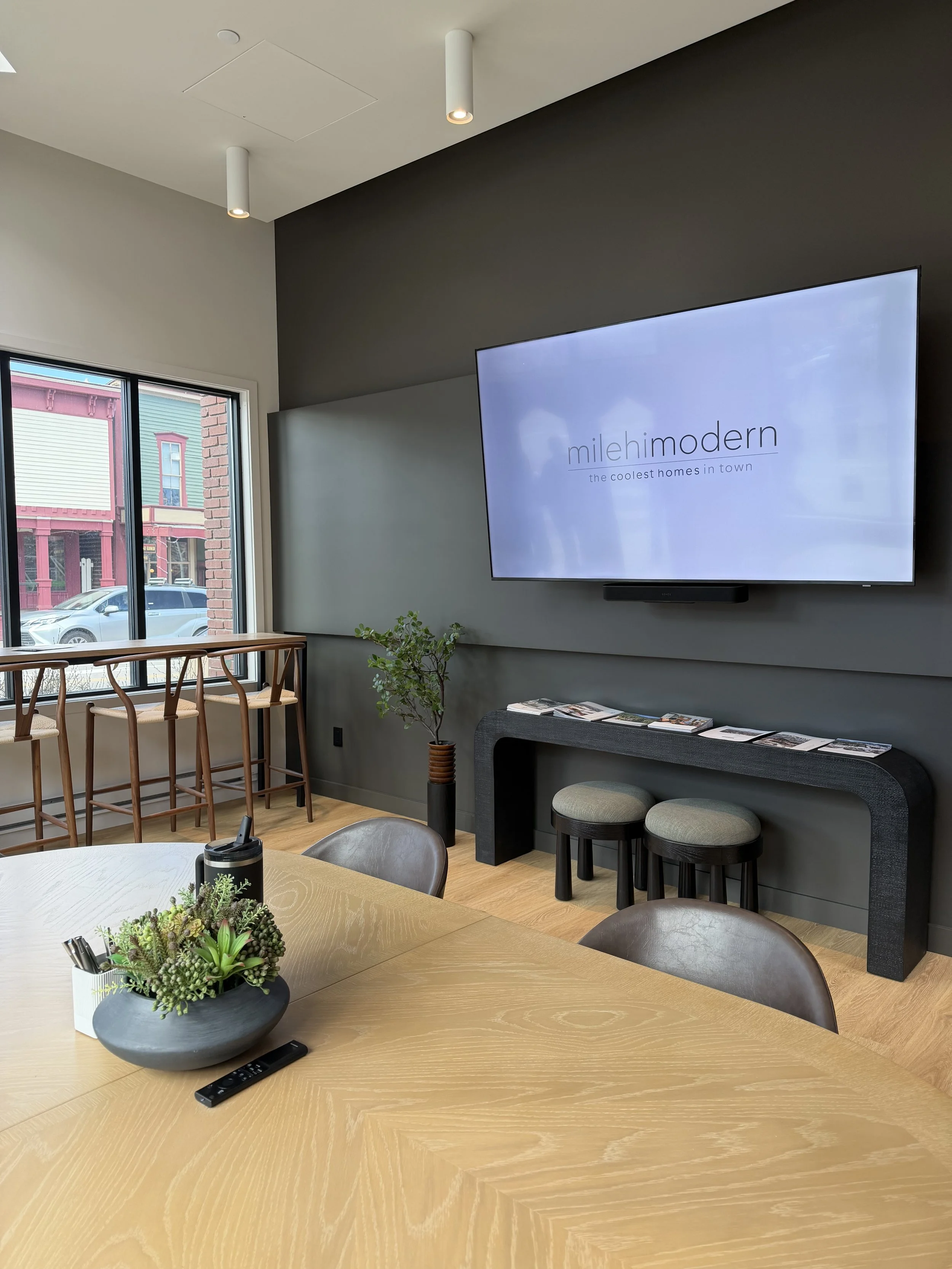 Interior of a modern office or conference room with a large wooden table, a potted plant, a TV screen on the wall displaying 'mileh modern, the coolest homes in town,' a window with a street view, and bar stools along the window.