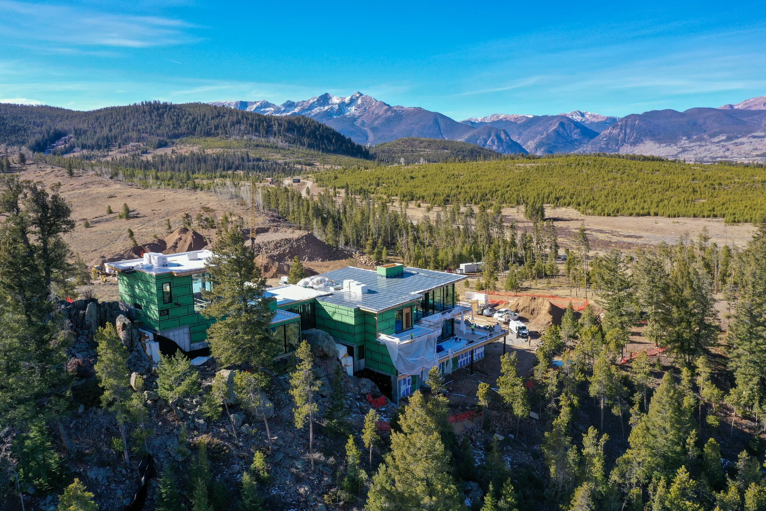 A residential construction site in a forested mountainous area with a house under construction, surrounded by trees and mountain views in the background.