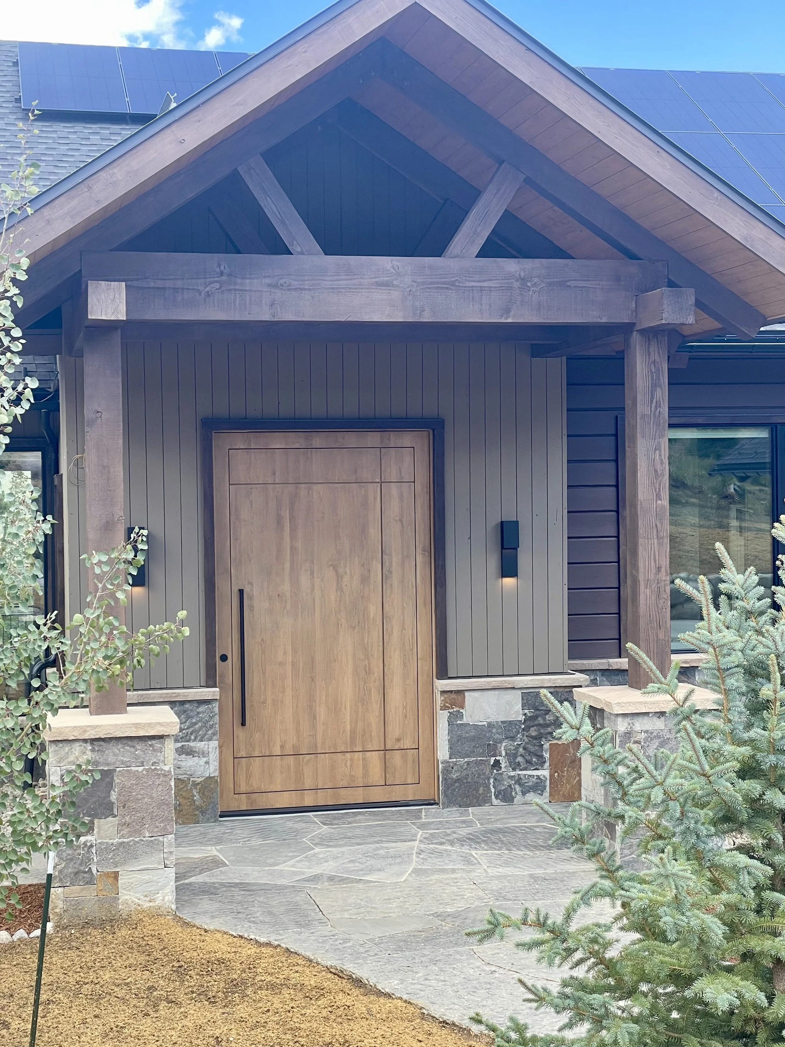 A modern house entrance with a wooden door, stone foundation, wood accents, black vertical siding, and solar panels on the roof. Vegetation and a pathway are visible in the foreground.