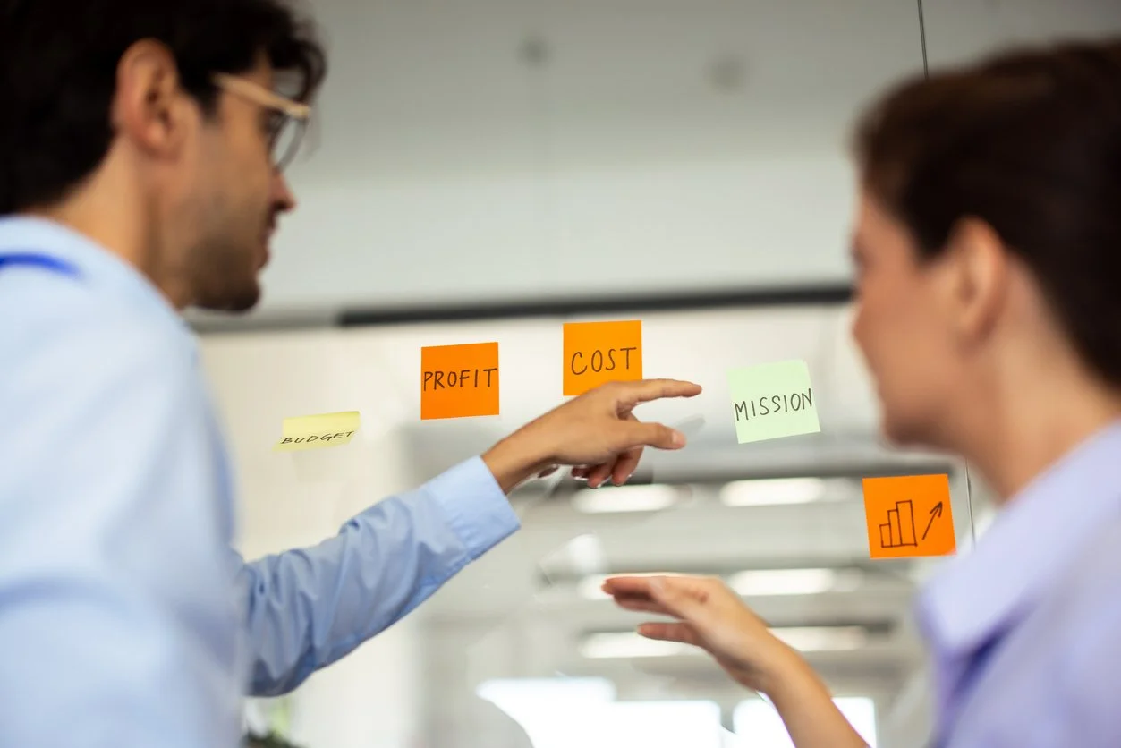 a man in business attire pointing at a yellow sticky note that says mission. there is a women standing next to him in business attire. next to the mission sticky noters are the sticky notes budget, profit, cost, and a bar graph with an up arrow