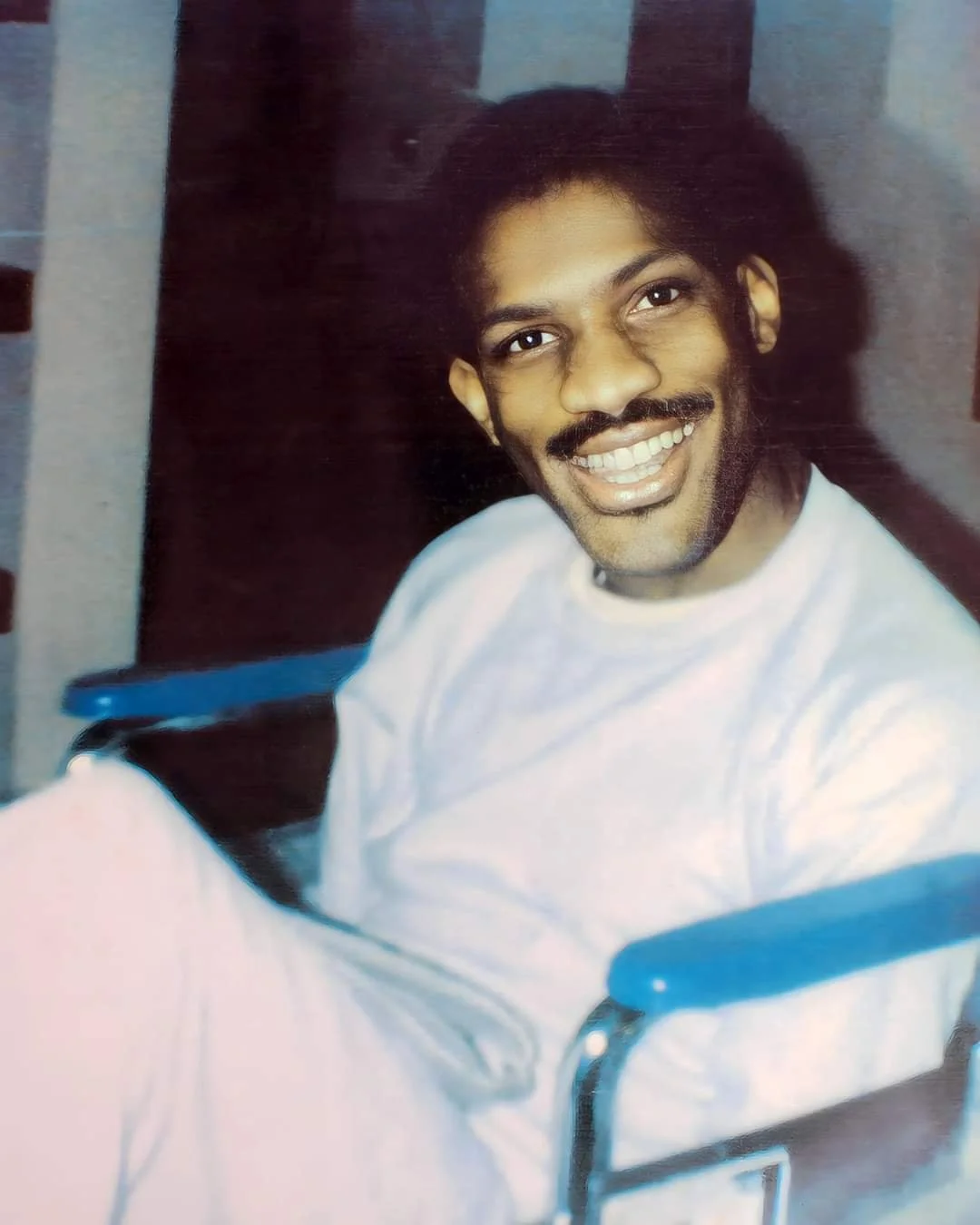 A smiling man with dark, curly hair and a mustache, wearing a white shirt, sitting in a wheelchair indoors.