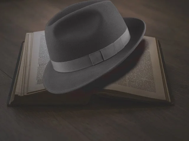 A gray fedora hat with a ribbon band resting on an open book on a wood surface.