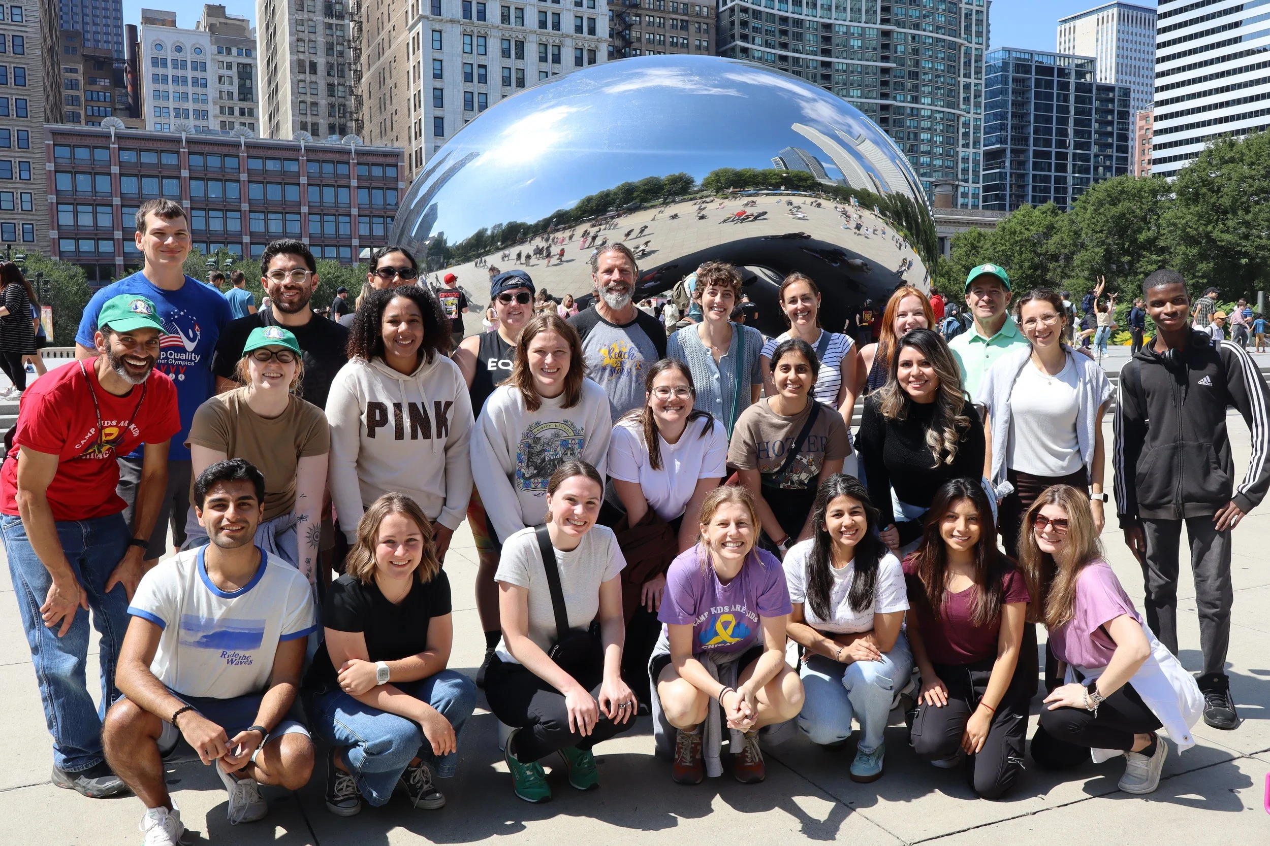 A group of diverse people posing in front of the Cloud Gate sculpture in Millennium Park, Chicago, on a sunny day with city buildings in the background.