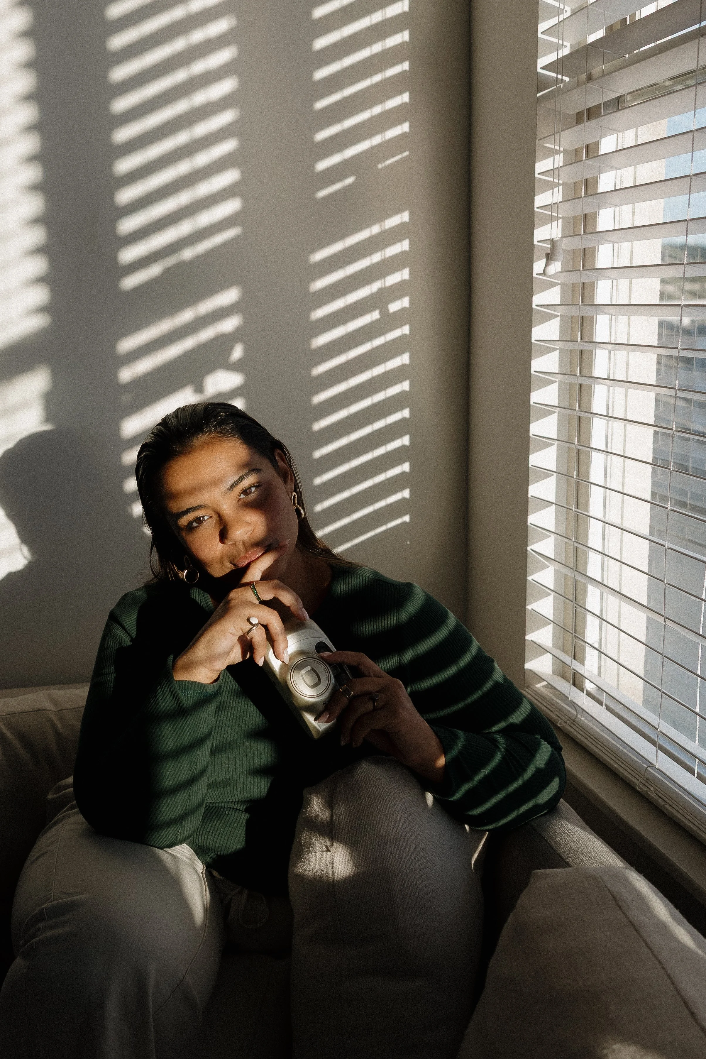 Mexico Wedding Photographer A woman sitting on a sofa near sunlit window blinds, holding a vintage camera, looking at the camera with a thoughtful expression.