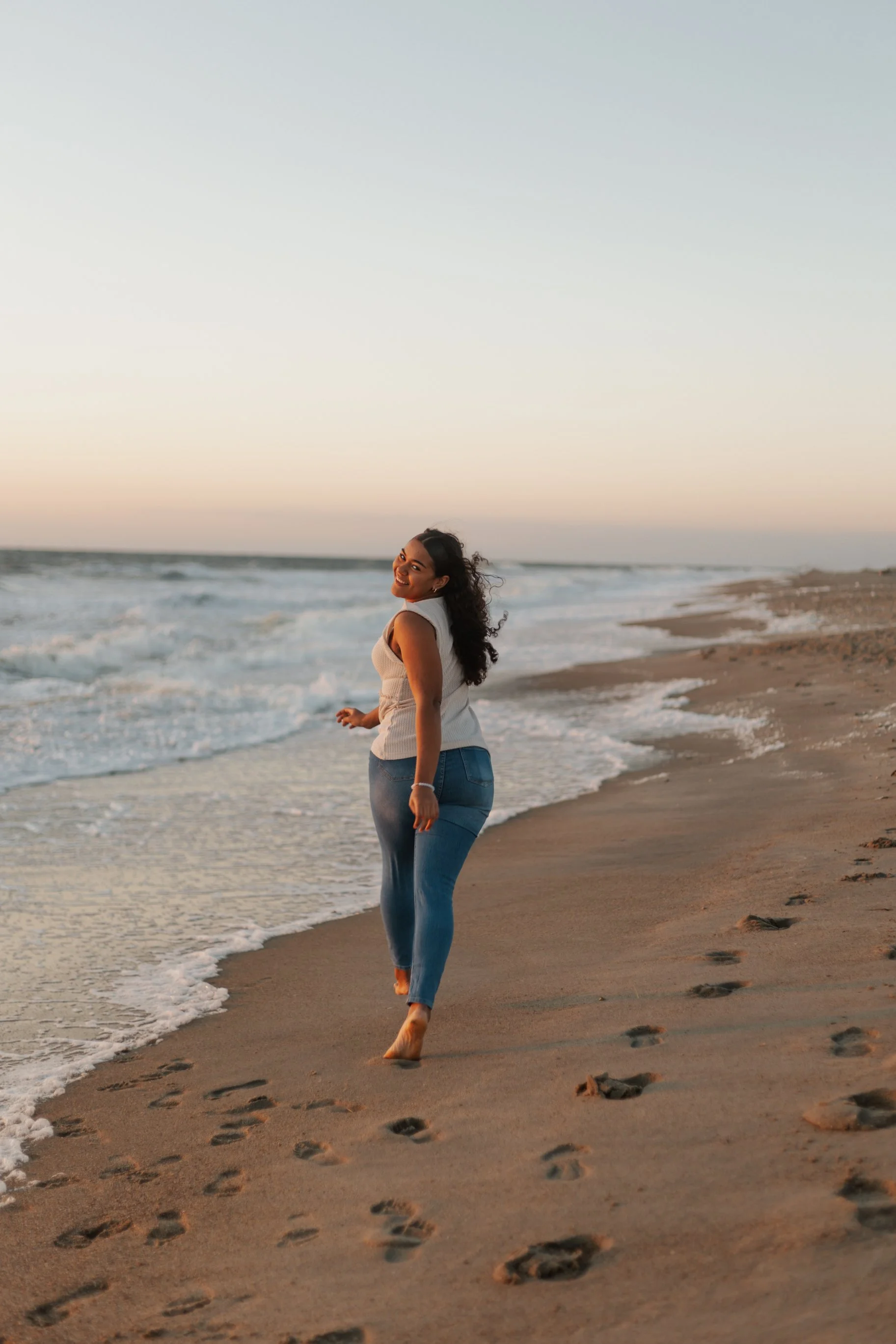 A woman with curly dark hair walking barefoot on a sandy beach near the ocean at sunset, smiling happily.