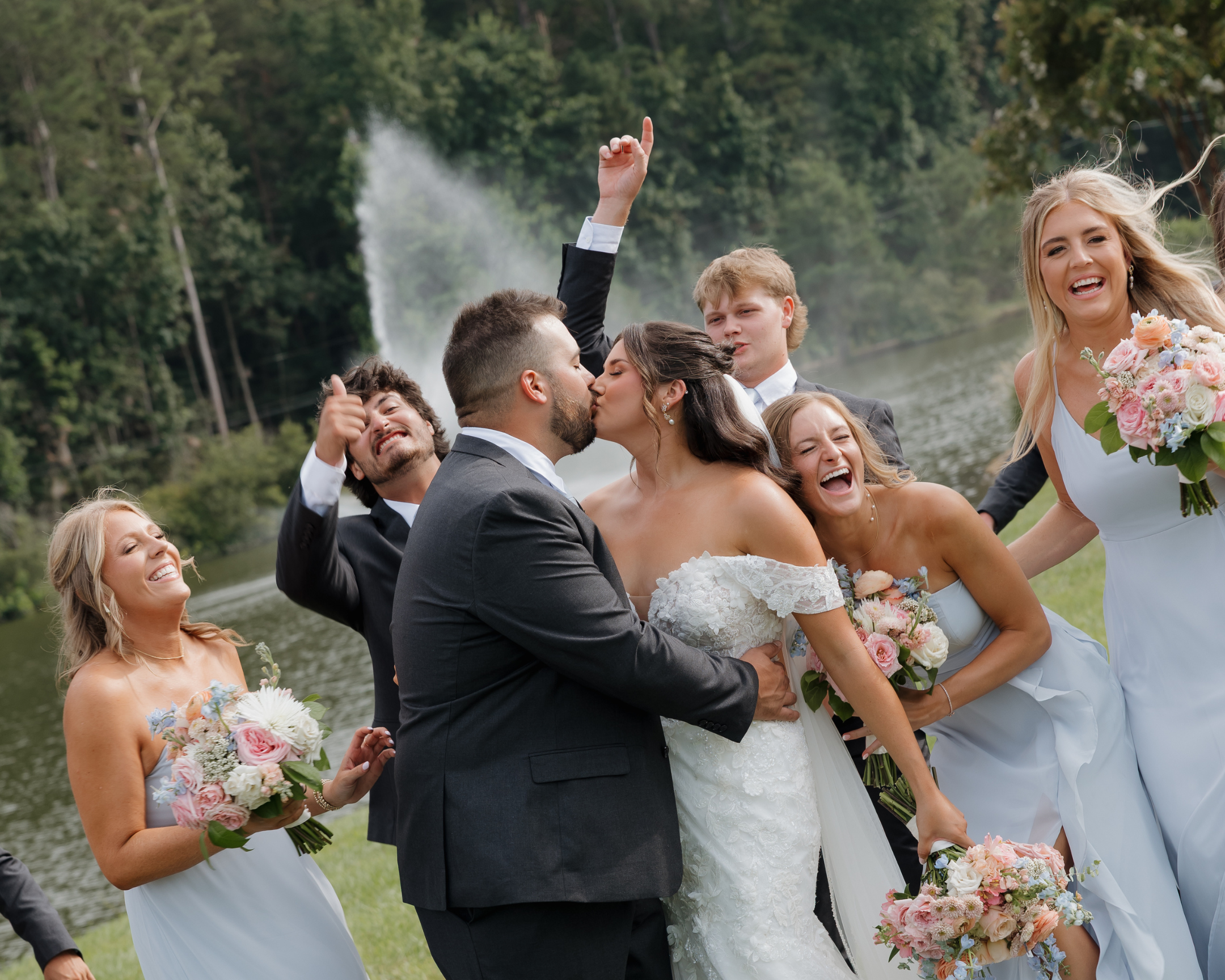 DC Wedding Photographer. Michaela Mae A group of wedding guests celebrating outdoors near a river, with a waterfall in the background. A bride and groom are kissing, surrounded by bridesmaids and groomsmen holding bouquets and making joyful gestures.