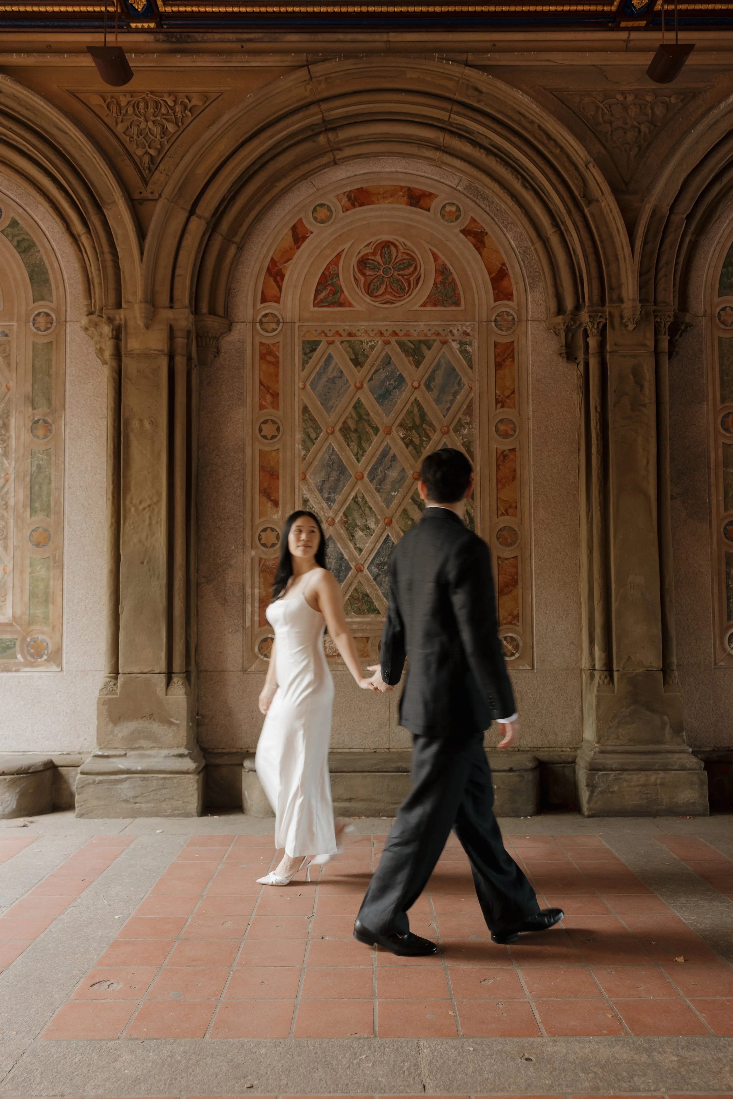 DC Wedding Photographer. A couple holding hands, walking in front of an ornate, stained glass and stone archway. The woman wears a white dress and heels, the man a dark suit.