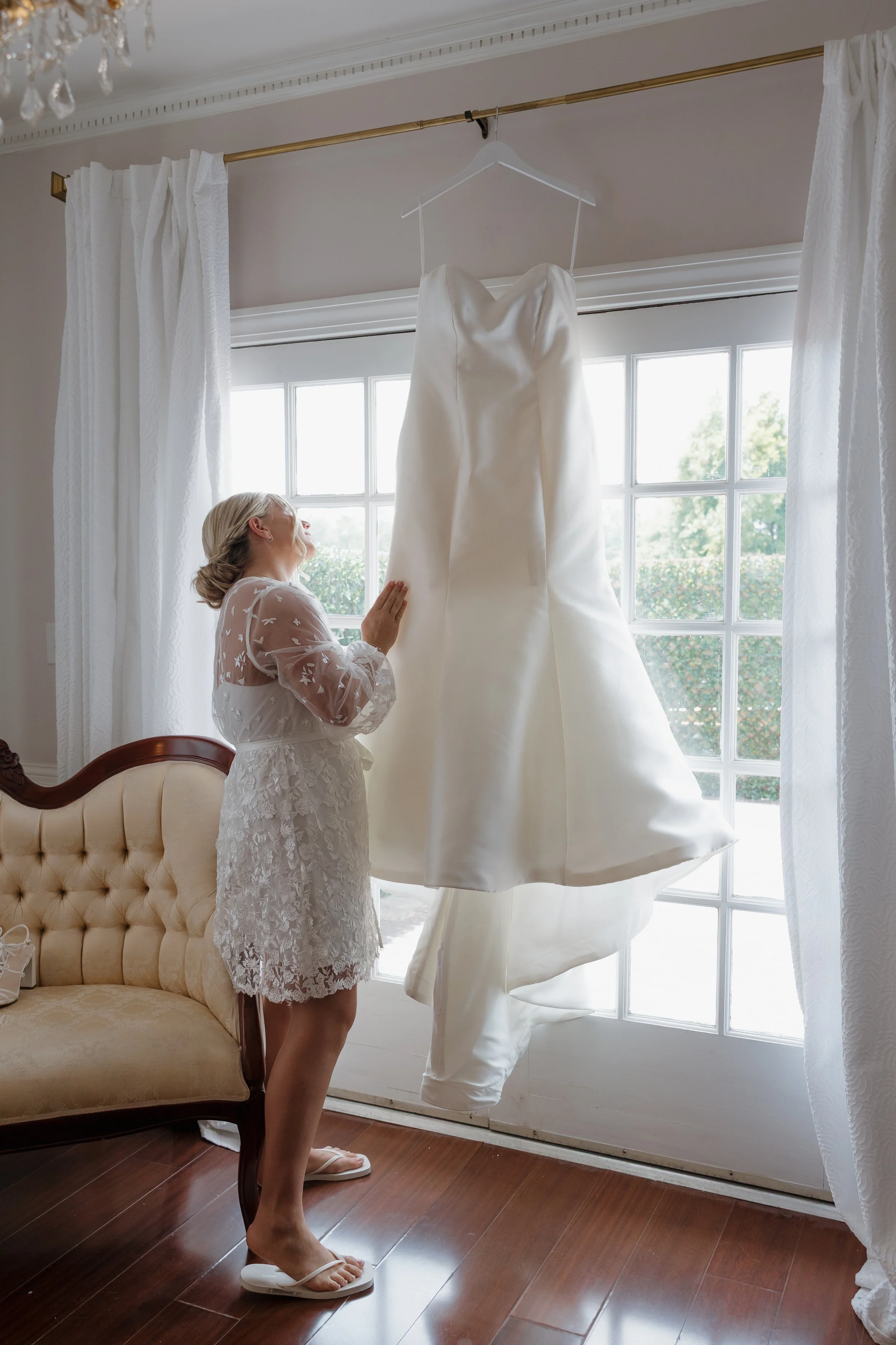 DC Wedding Photographer. Michaela Mae and Co. A woman in a lace robe stands next to a beige vintage style sofa, looking at a hanging white wedding dress in front of a large window with white curtains, with trees visible outside.