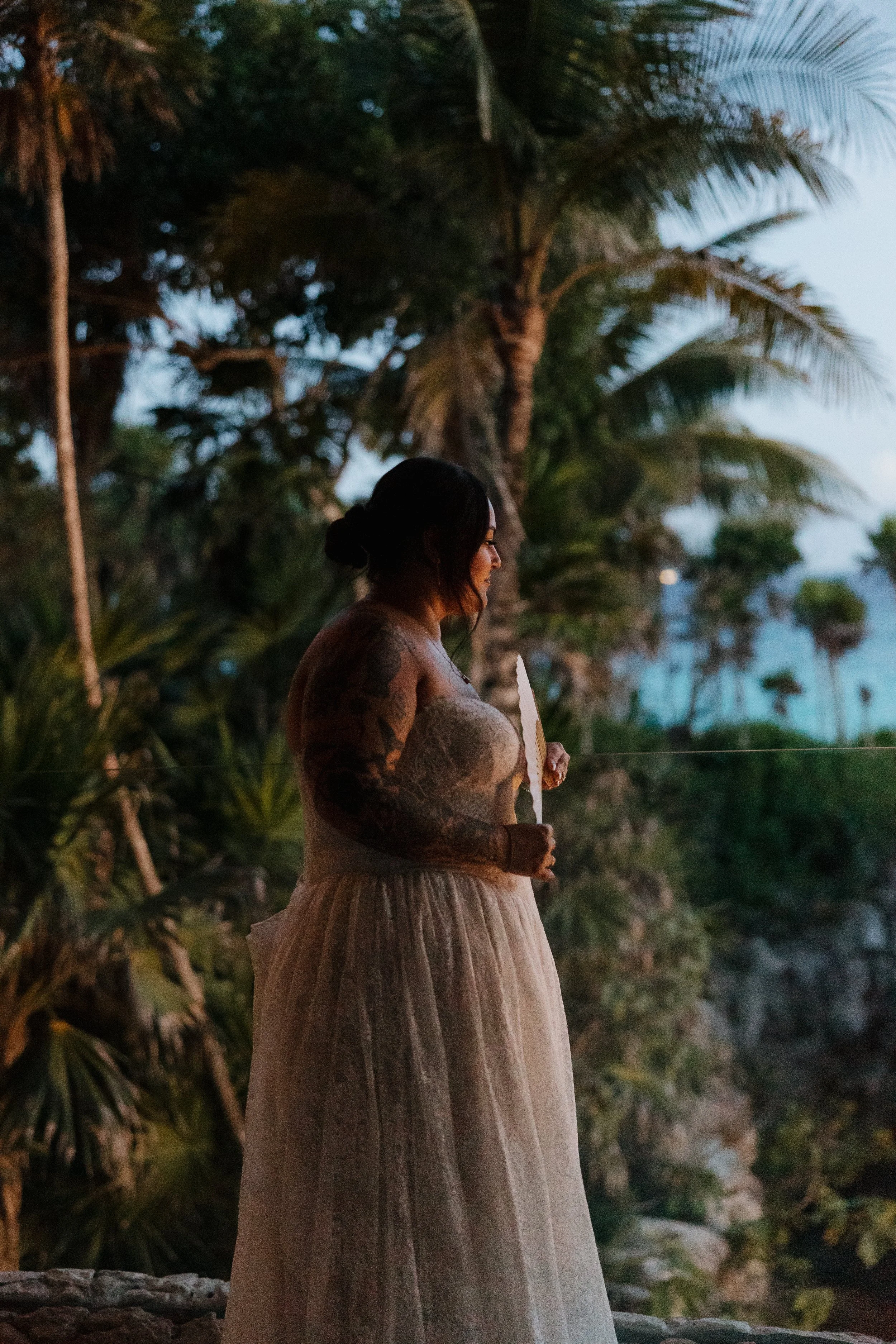 Mexico Wedding Photographer A woman with tattoos wearing a dress holding a fan, standing outdoors during sunset with palm trees and a body of water in the background.