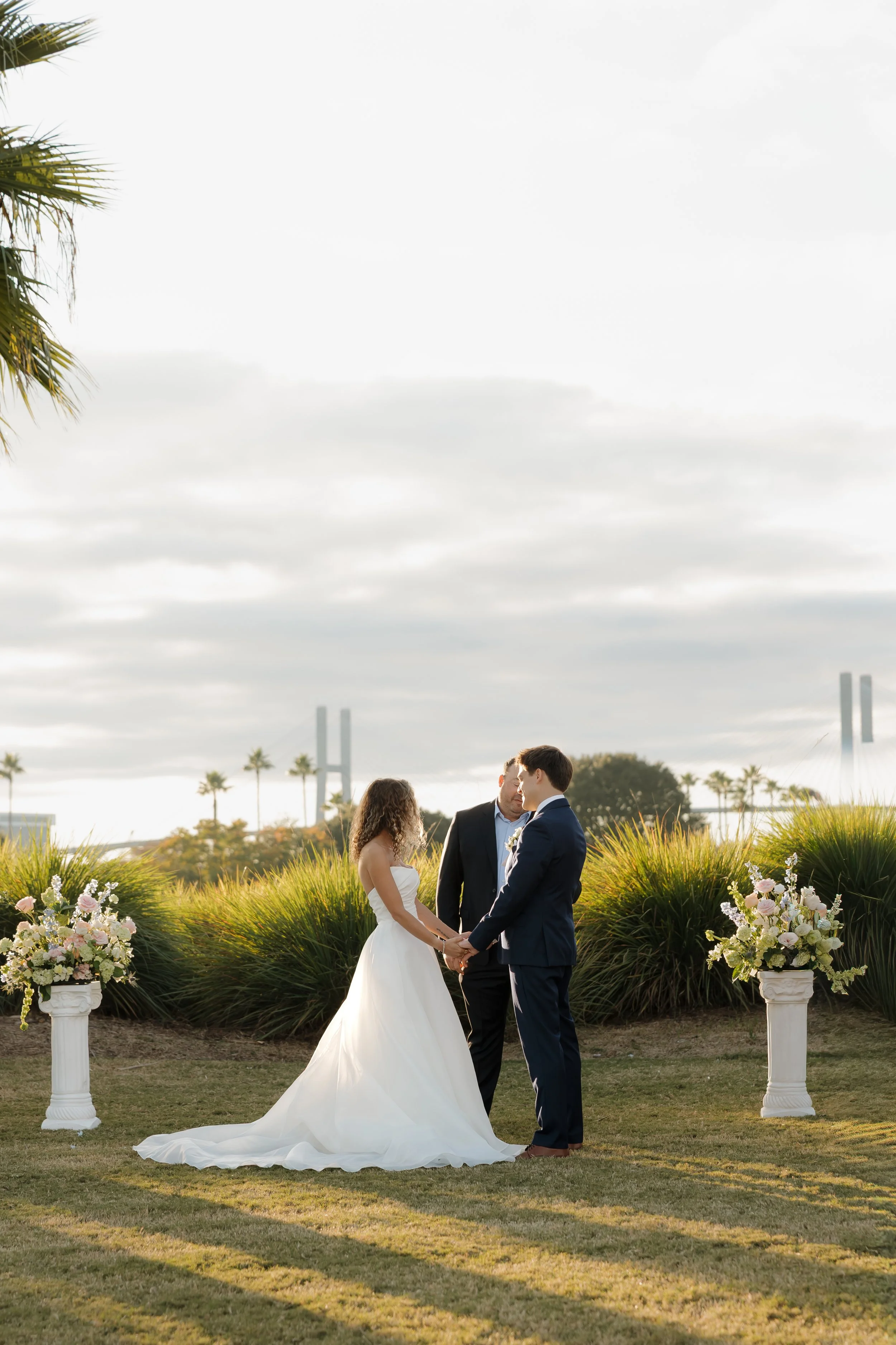 DC Wedding Photographer. Michaela Mae and Co. A wedding ceremony outdoors at sunset with a bride and groom holding hands and a officiant. Floral arrangements in white urns are on either side of them, and palm trees and a bridge are in the background.
