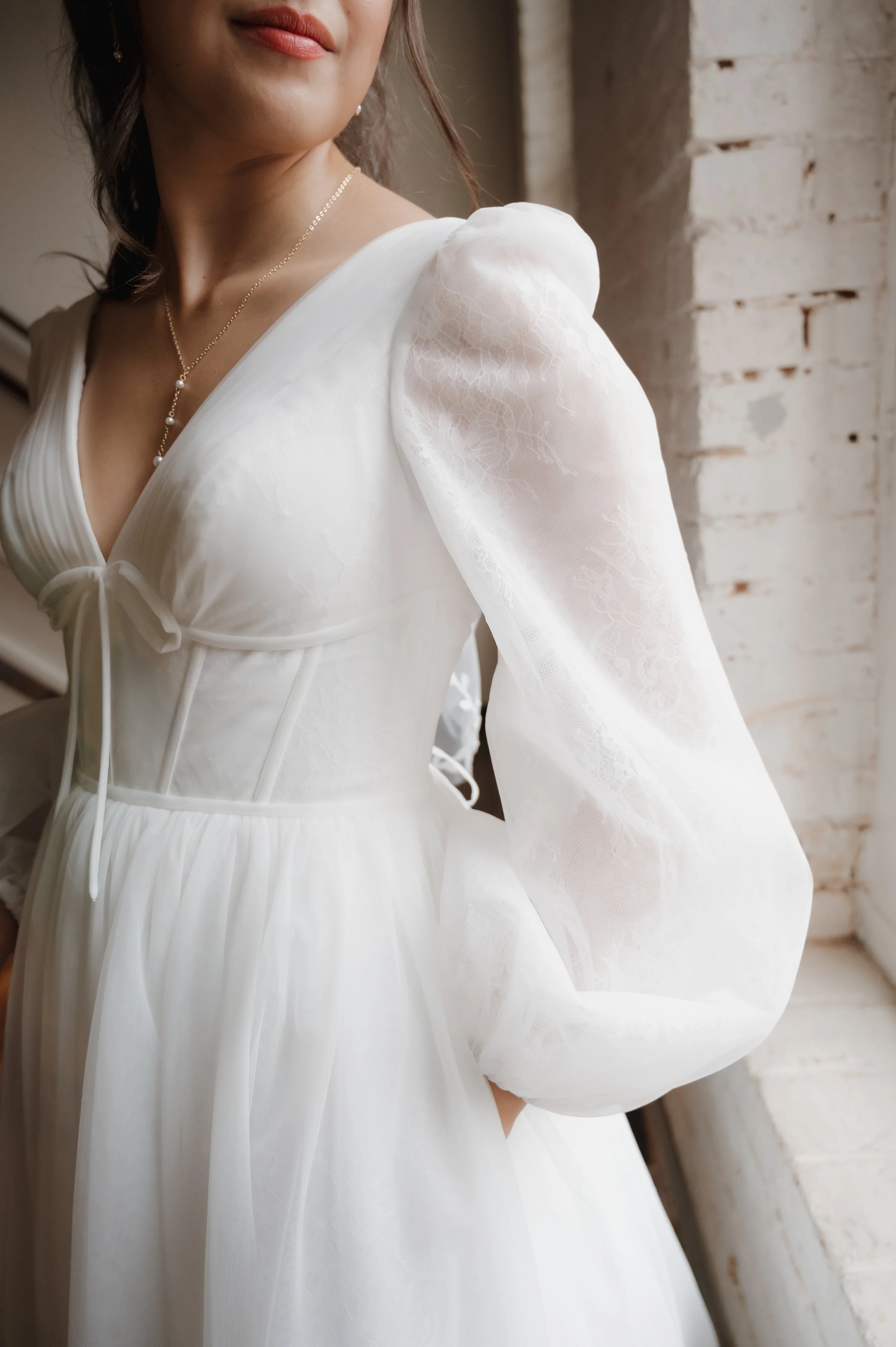 DC Wedding Photographer  Close-up of a woman wearing a white dress with puffed lace sleeves, standing near a window with a brick wall background.
