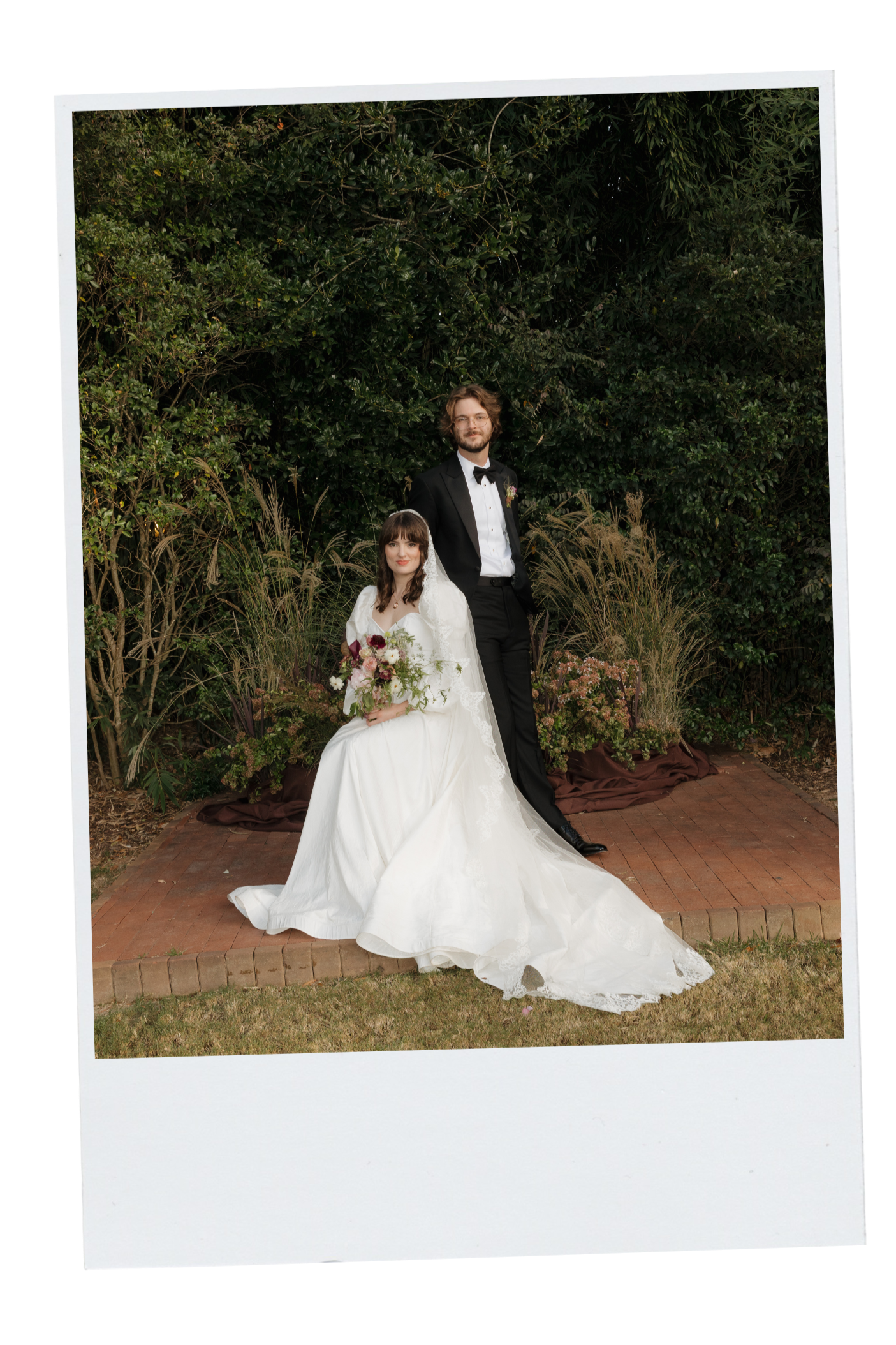 A bride and groom posing for a wedding photo outdoors, with greenery background. The bride is seated in a white wedding dress holding a bouquet, and the groom stands beside her in a black tuxedo. DC Wedding Photographer