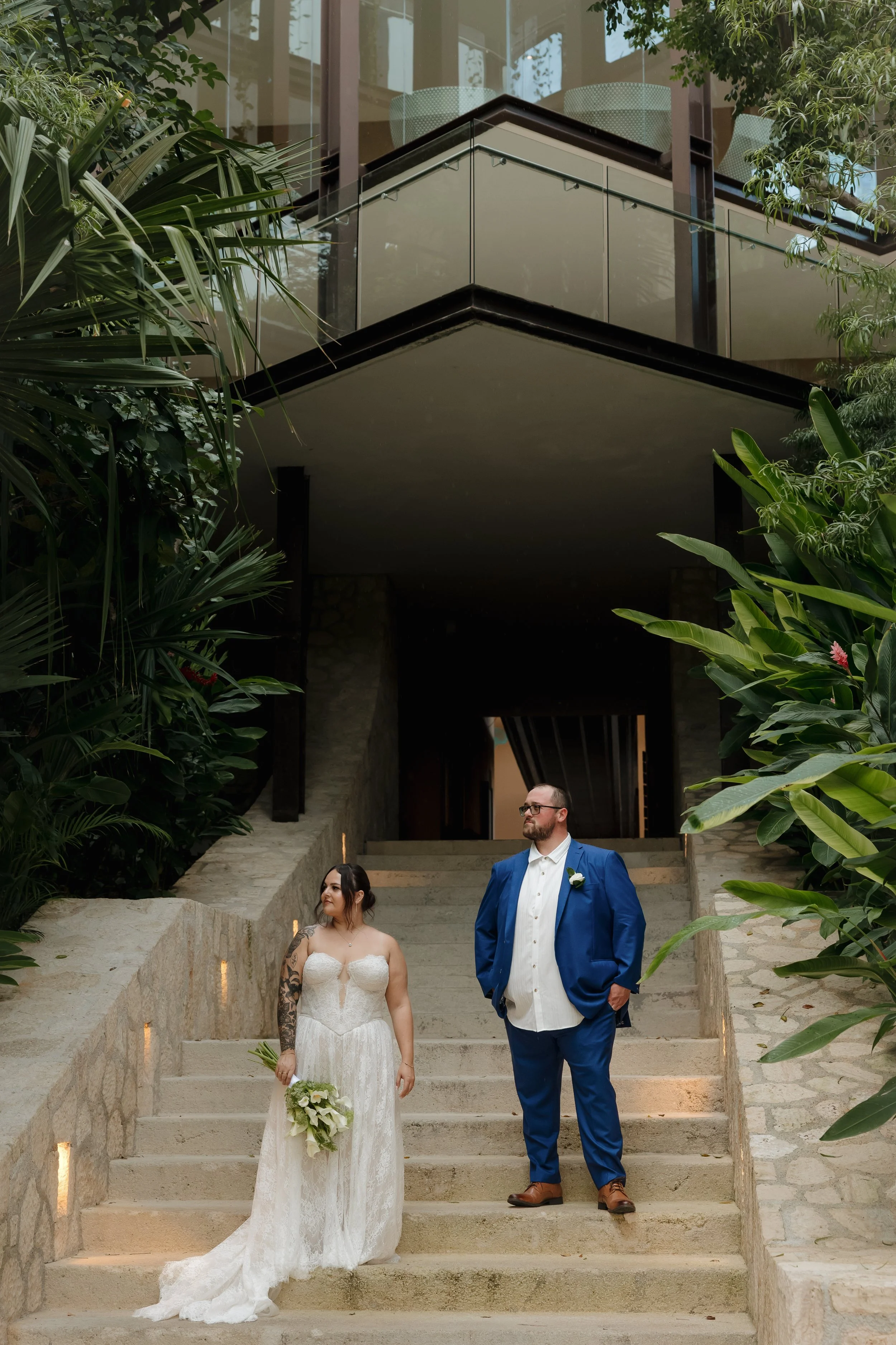 Mexico Wedding Photographer A bride in a white wedding gown holding a bouquet of white flowers and a groom in a blue suit standing on stone steps surrounded by greenery.