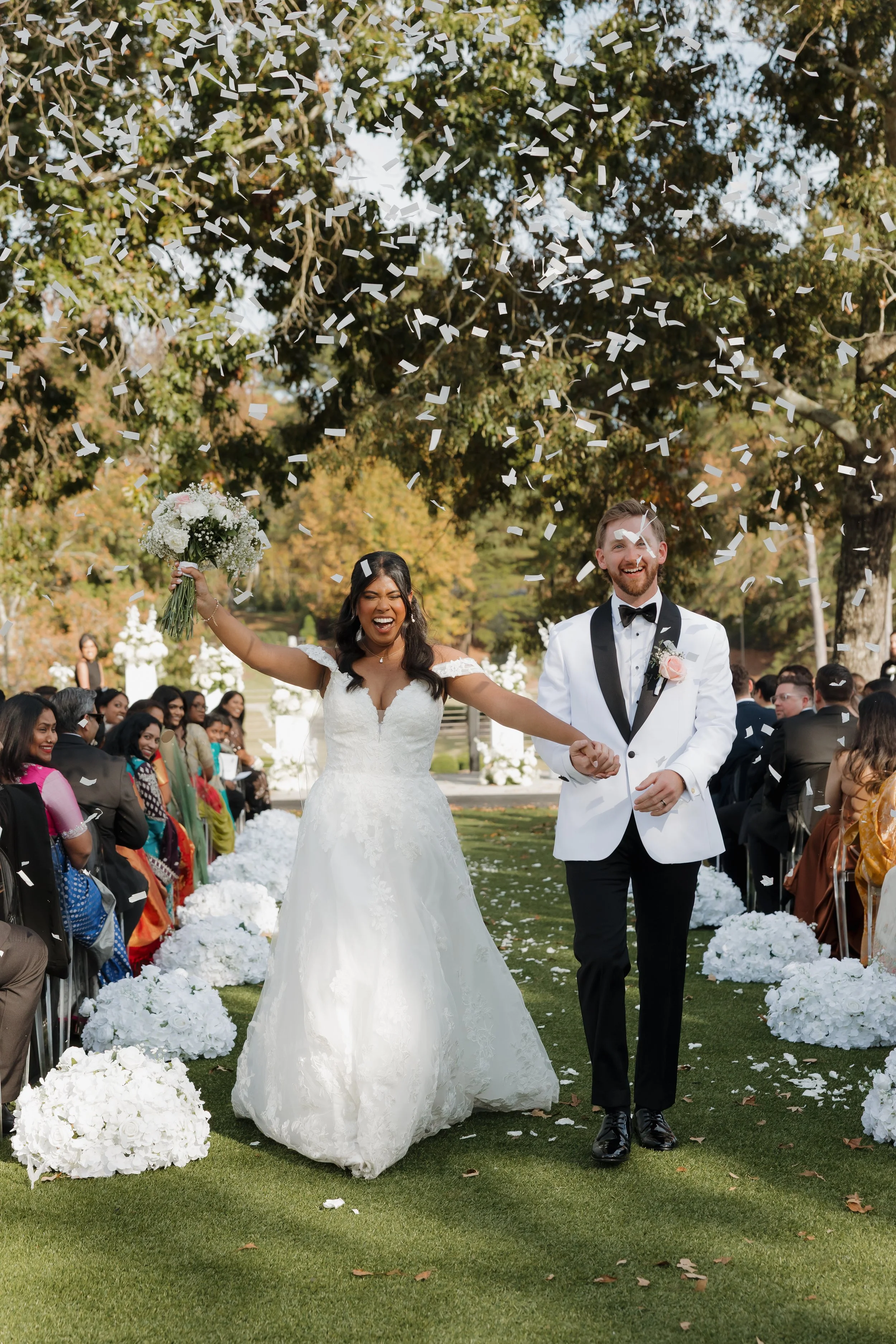 DC newlywed couple, the bride in a white wedding dress holding a bouquet, and the groom in a white tuxedo jacket with black pants, celebrating as confetti falls from above during their wedding ceremony outdoors with guests seated on either side.