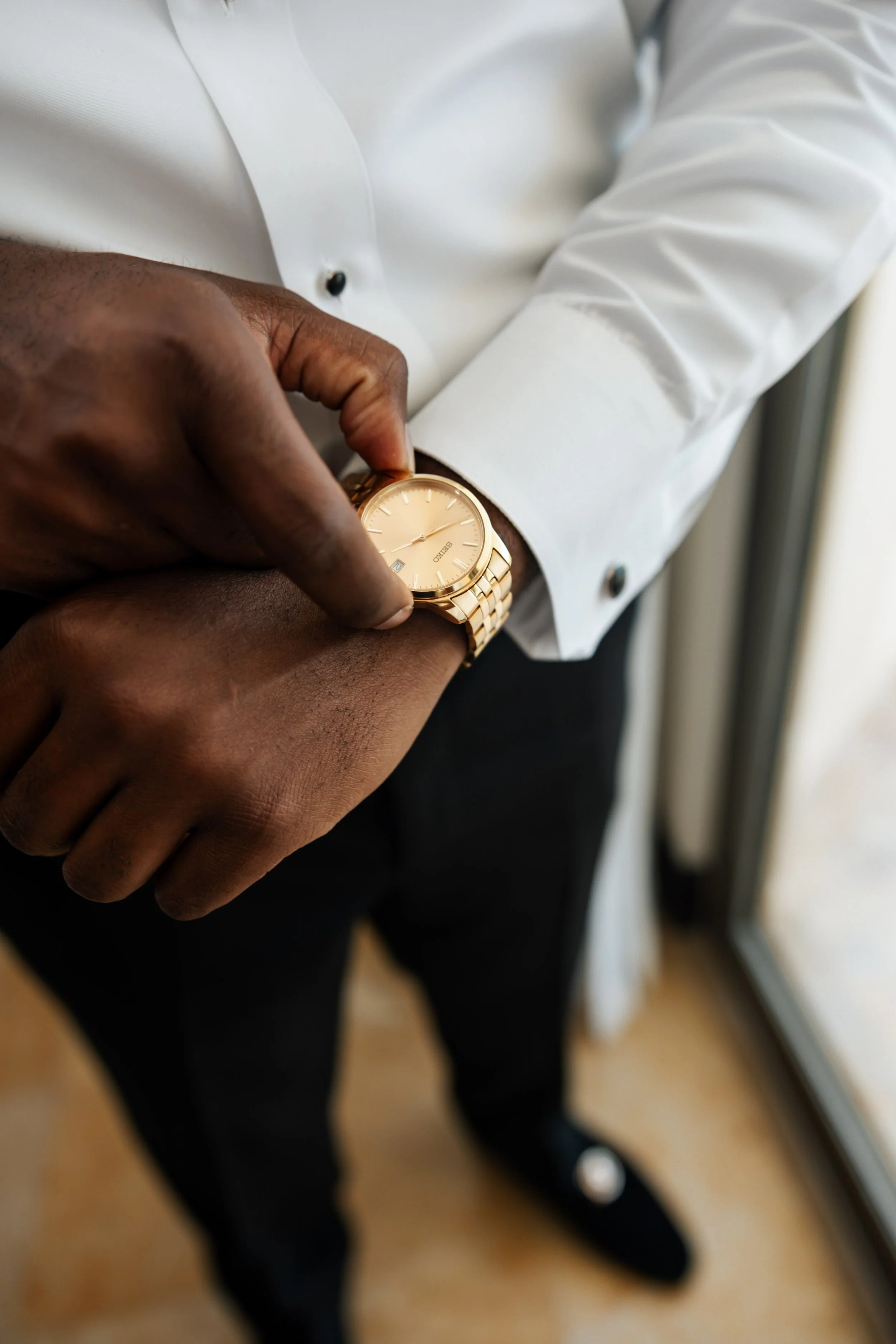 Editorial Washington DC Wedding Photos A man adjusting a gold wristwatch while wearing a white dress shirt and black pants, standing indoors near a window.
