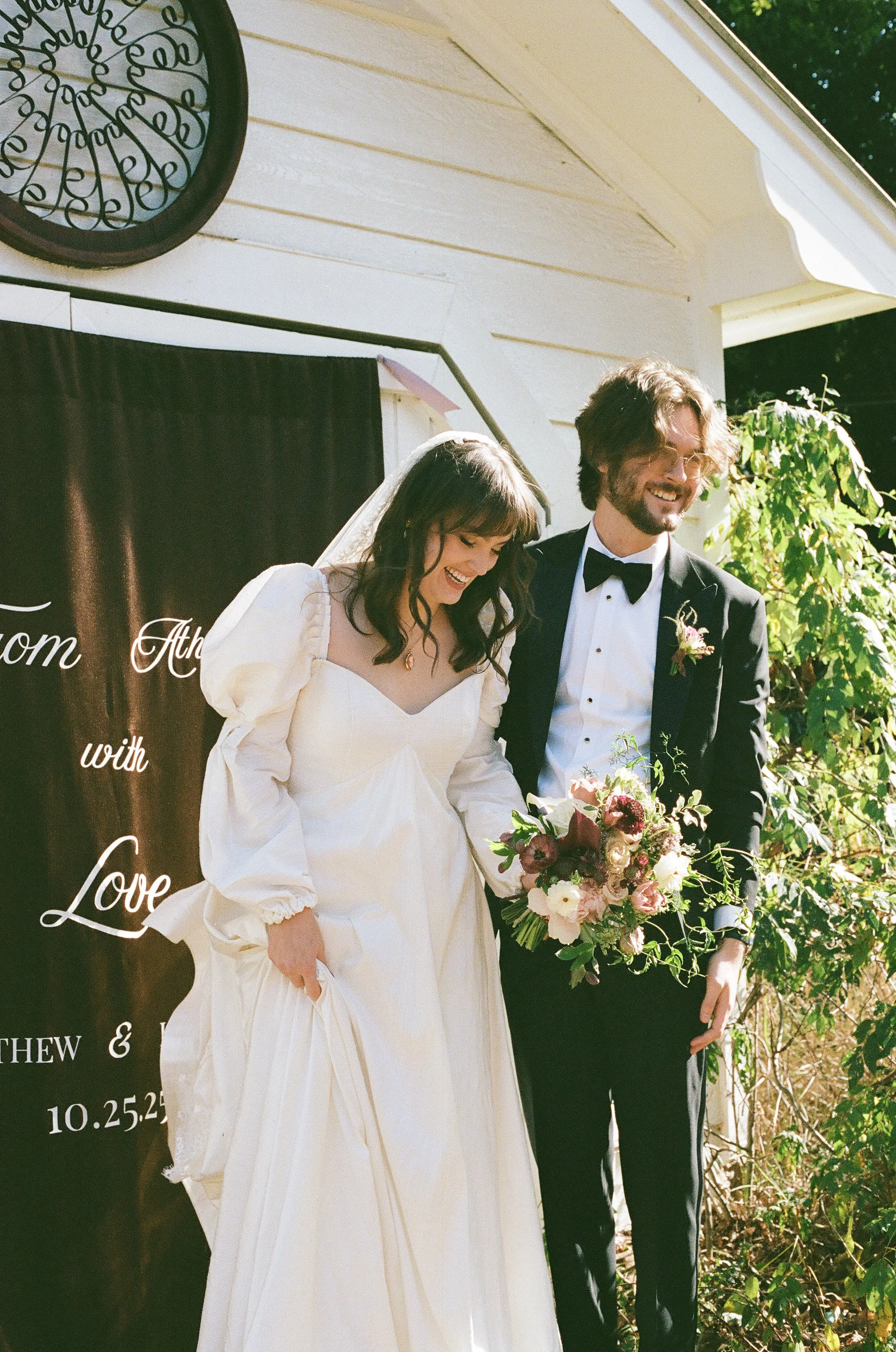 DC Wedding Photographer. Michaela Mae and Co. A bride in a white wedding dress and a groom in a black tuxedo celebrate outdoors, with the bride holding a bouquet of flowers, in front of a black banner with white text.