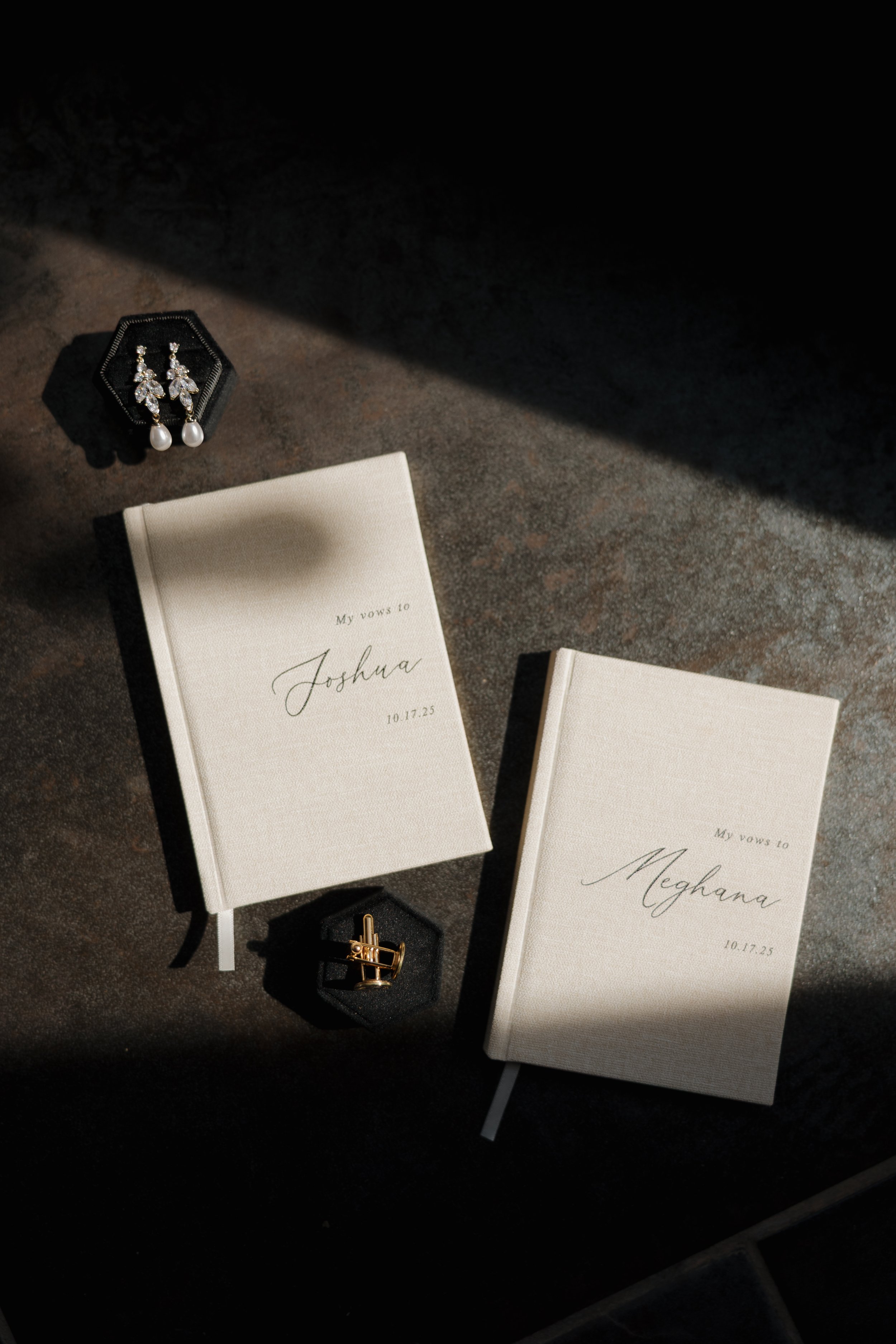 DC Wedding Photographer. Michaela Mae and Co. Two white vow books with names Joshua and Meghana, dated 10.17.25, a pair of pearl and diamond earrings, and a gold ring displayed on a dark surface with a shadow across the scene.