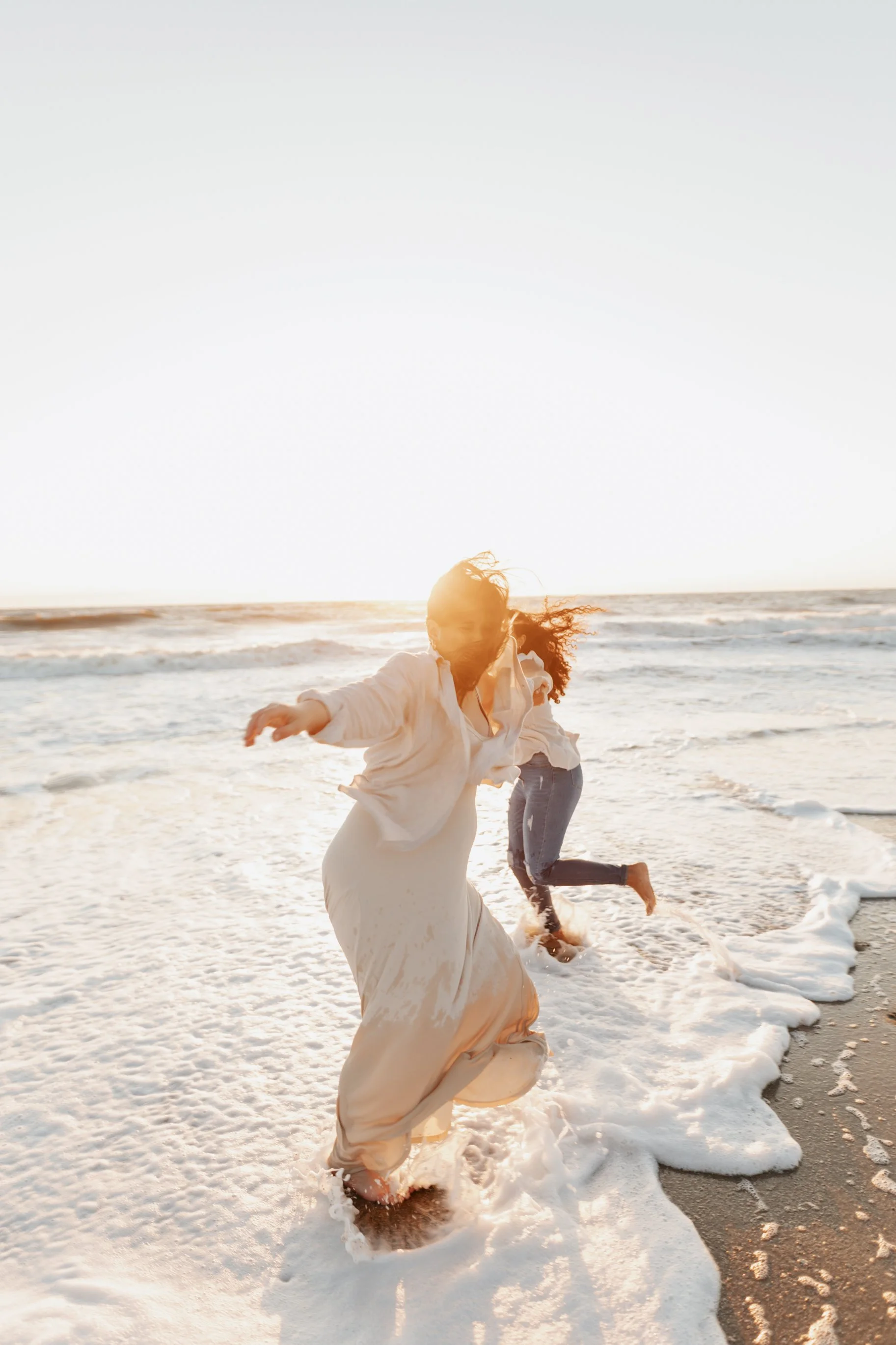 Mom and child playing and running on the beach at sunset, with waves and foam around their feet.