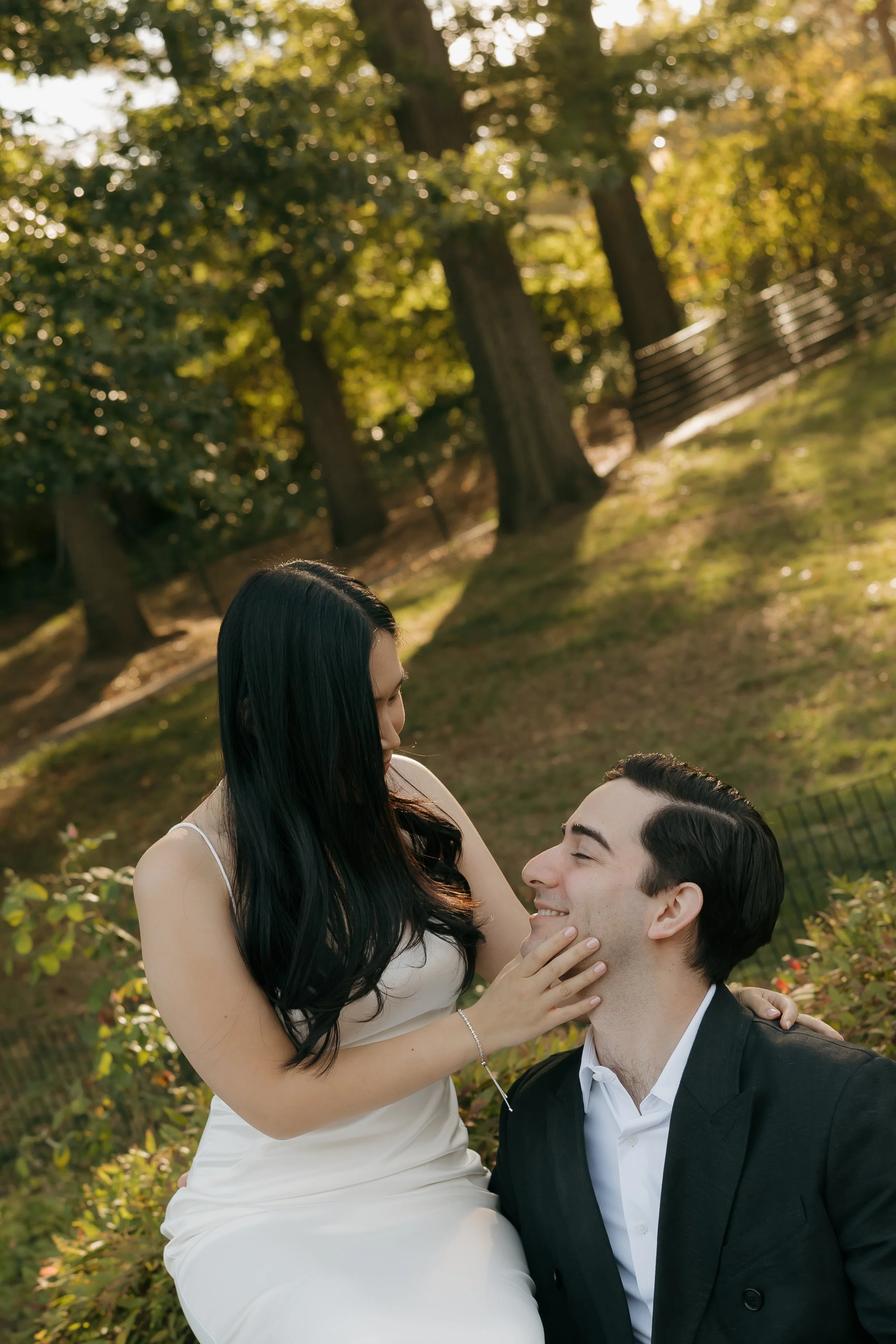 DC Wedding Photographer. A couple sharing a romantic moment outdoors in a park, with the woman gently touching the man's face as they smile at each other, surrounded by green trees and sunlight.
