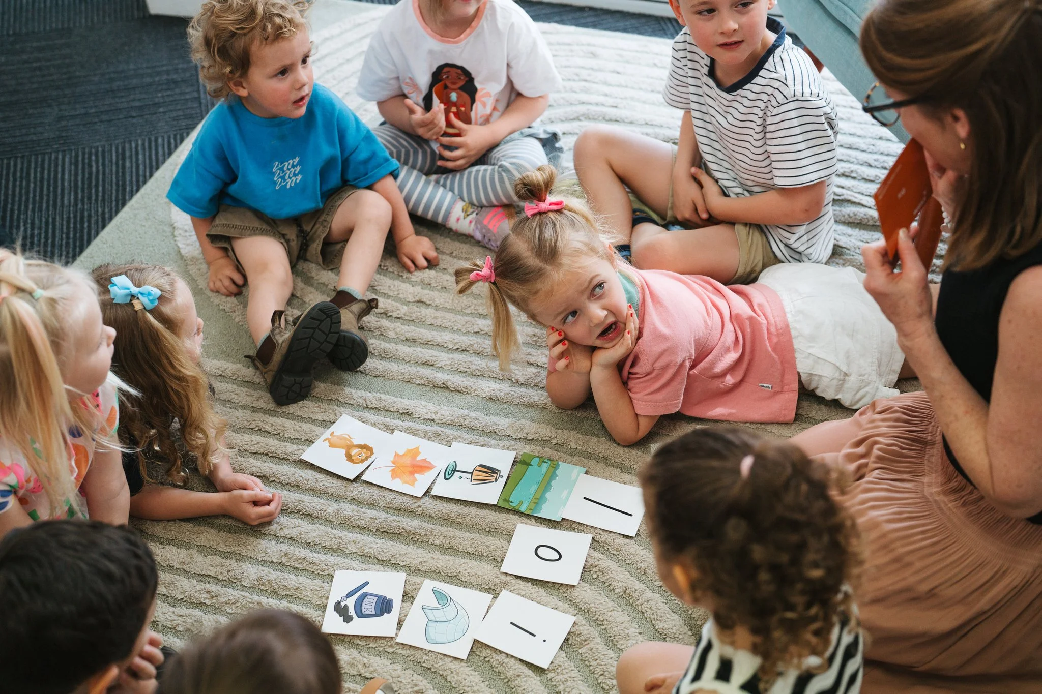 A woman reading a picture book about koalas to a group of five children outdoors on a sunny day. The children are sitting on the grass near a large tree with a white trunk, surrounded by rocks and greenery.