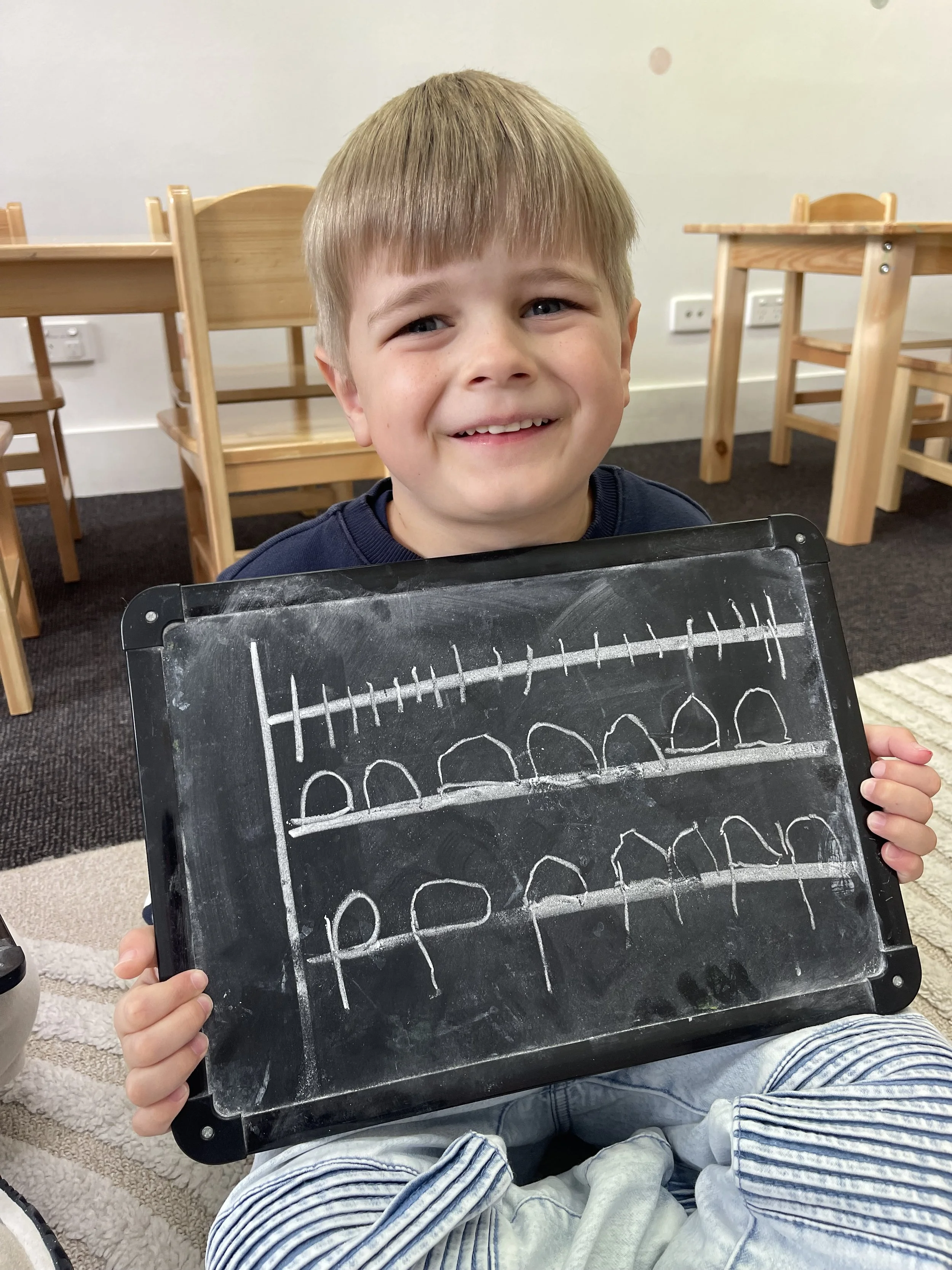 A young girl with blonde hair and a large white bow in her hair is writing on a laminated sheet with red, yellow, and blue borders, practicing the phrase "A DAY AT" and drawing shapes in blue marker.
