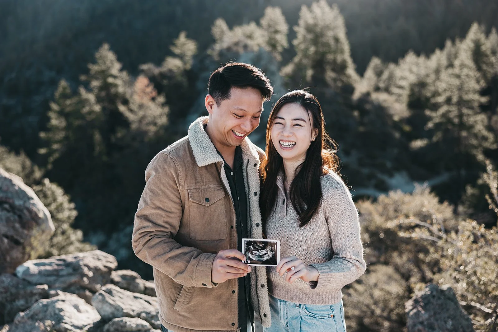 A man and a woman standing outdoors in a rocky, mountainous area, smiling and looking at an ultrasound scan held by the man.