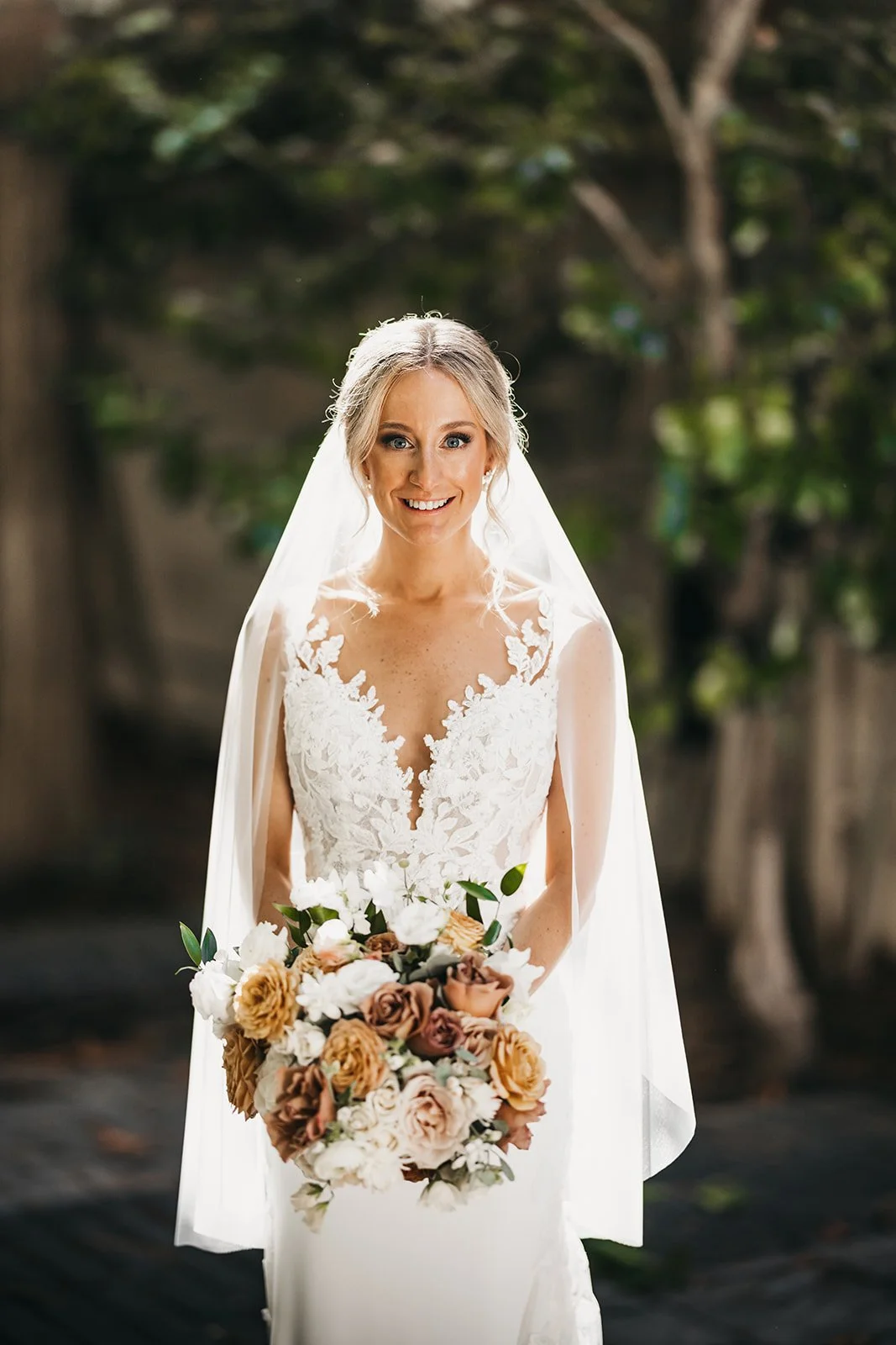 A smiling bride with blonde hair wearing a white lace wedding dress and veil, holding a bouquet of roses and other flowers outdoors with blurred trees in the background.