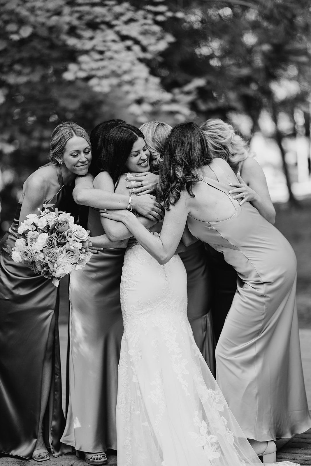 A group of women, including a bride in a white wedding gown, are hugging and celebrating outdoors, with trees in the background.