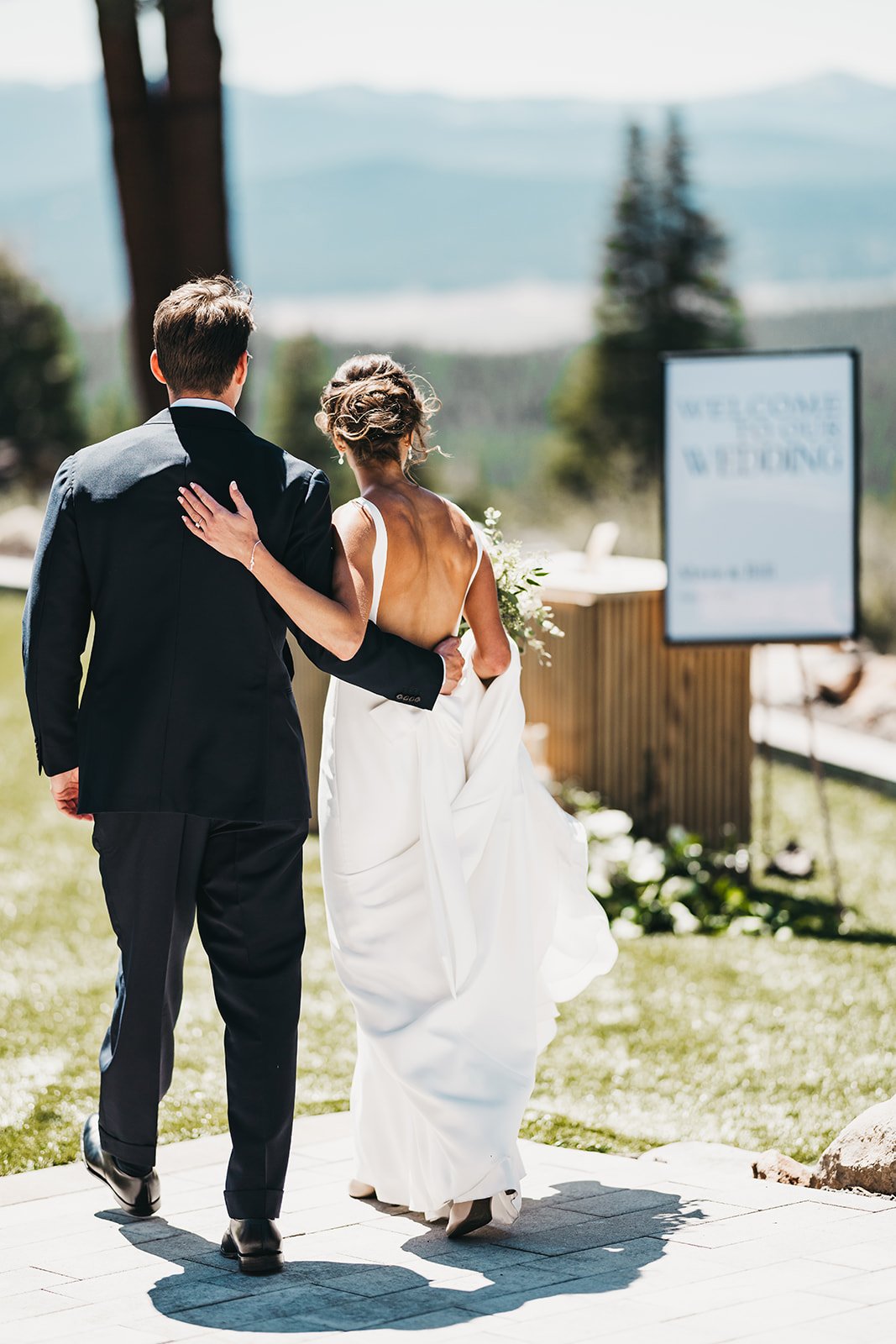 A couple walking arm-in-arm outdoors, the man in a dark suit and the woman in a white wedding gown, with a welcome sign and scenic mountain background at Austin Ridge, Truckee, CA.