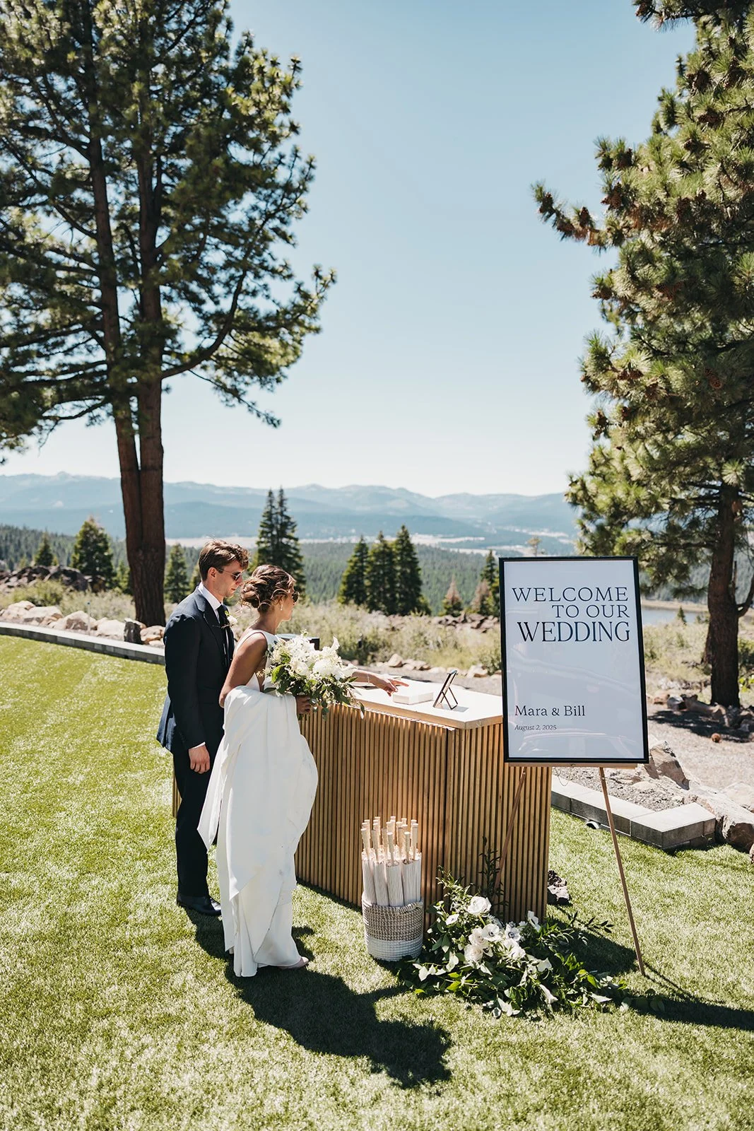 A bride and groom are signing documents during their outdoor wedding ceremony, with a sign that reads 'Welcome to Our Wedding Mara & Bill August 2, 2023' beside them, surrounded by trees and a scenic mountain landscape at Austin Ridge in Truckee, CA