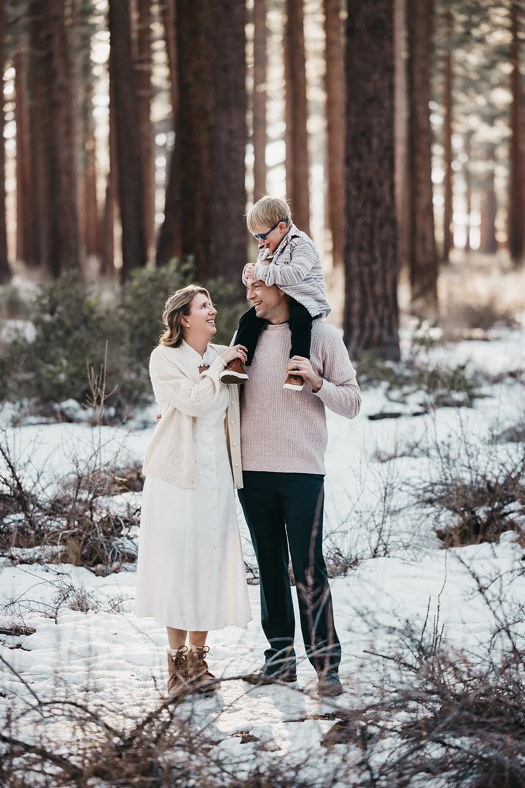 A family of three enjoying a walk in a snowy forest. The man is carrying a child on his shoulders, while the woman looks up at the child and smiles. All three are dressed warmly for winter.