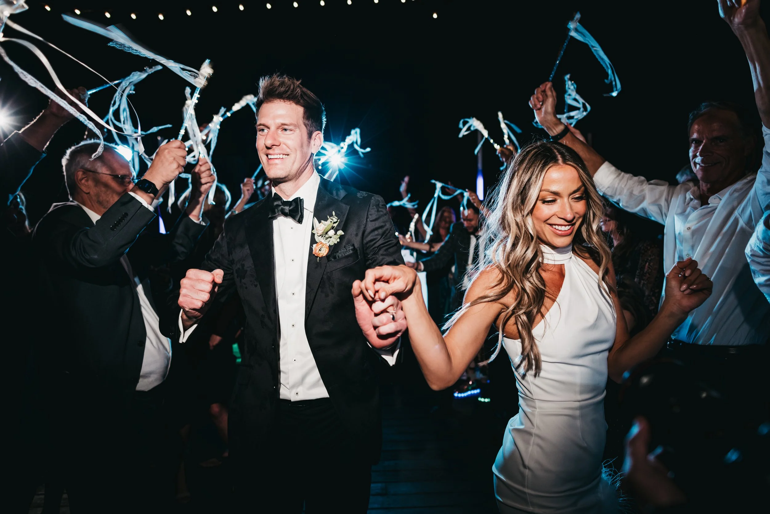 A newlywed couple, man in tuxedo and woman in white dress, dancing at their wedding reception, surrounded by guests celebrating with streamers and bright lights at Nakoma resort.
