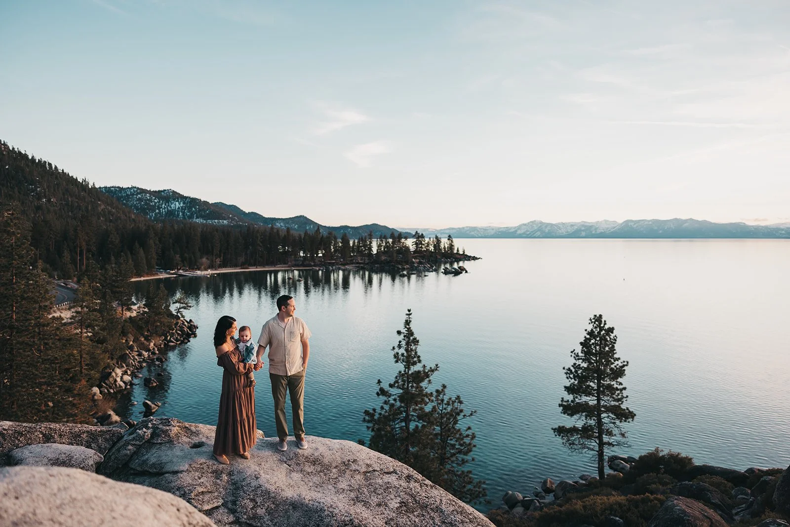A family of three standing on a large rock overlooking a lake surrounded by trees and mountains.