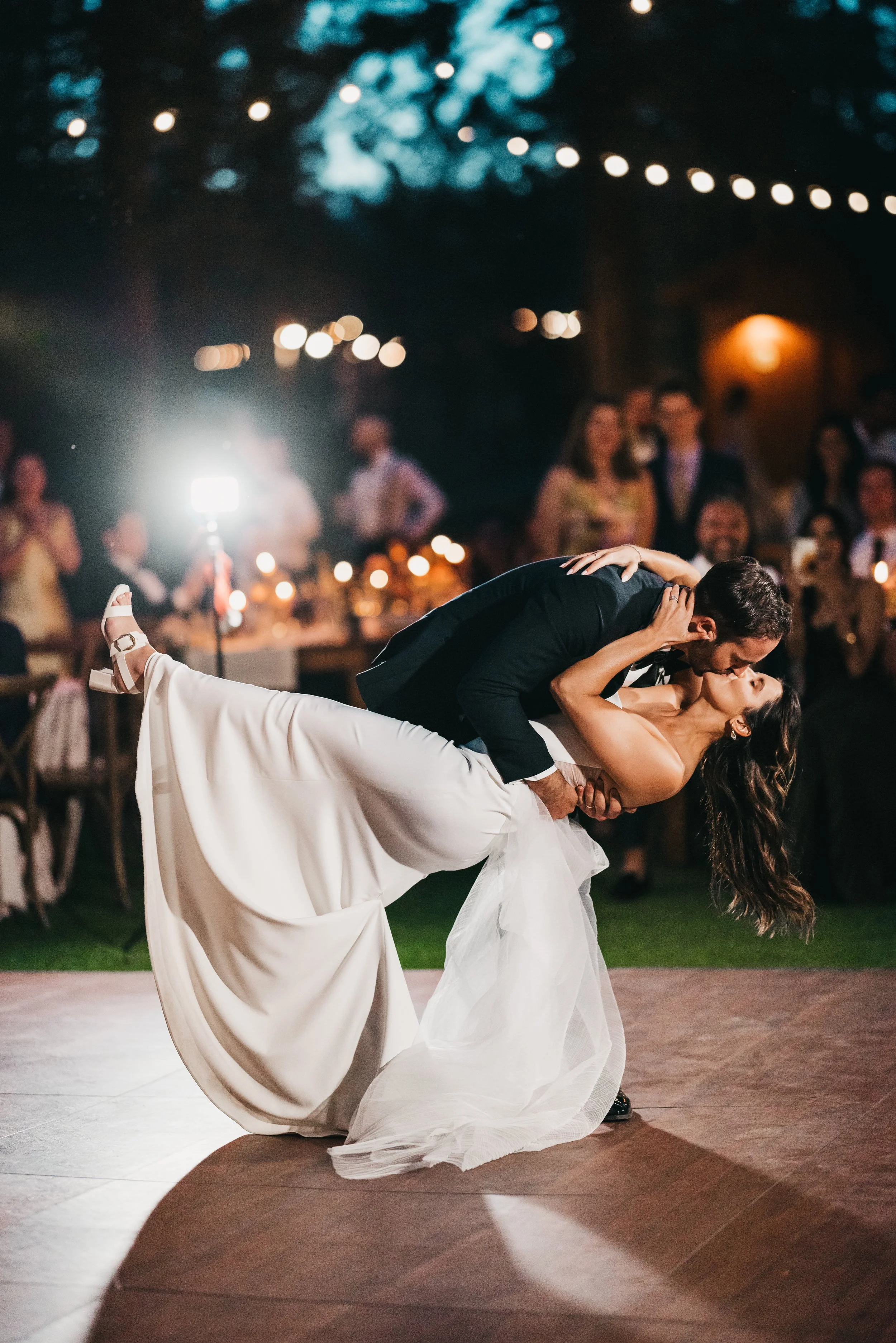 A newlywed couple dances on a wooden floor at their wedding reception, with the groom dipping the bride while sharing a kiss, surrounded by guests under string lights in an outdoor nighttime setting at Chalet View Lodge.