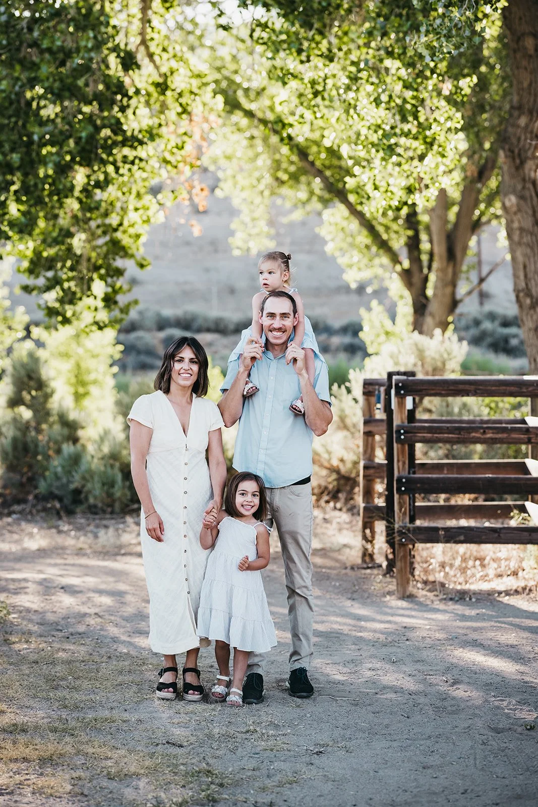 Family of four outdoors on a sunny day, with trees and a wooden fence in the background. The father is carrying a young girl on his shoulders, the mother is standing next to him holding the hand of a young girl, all smiling.