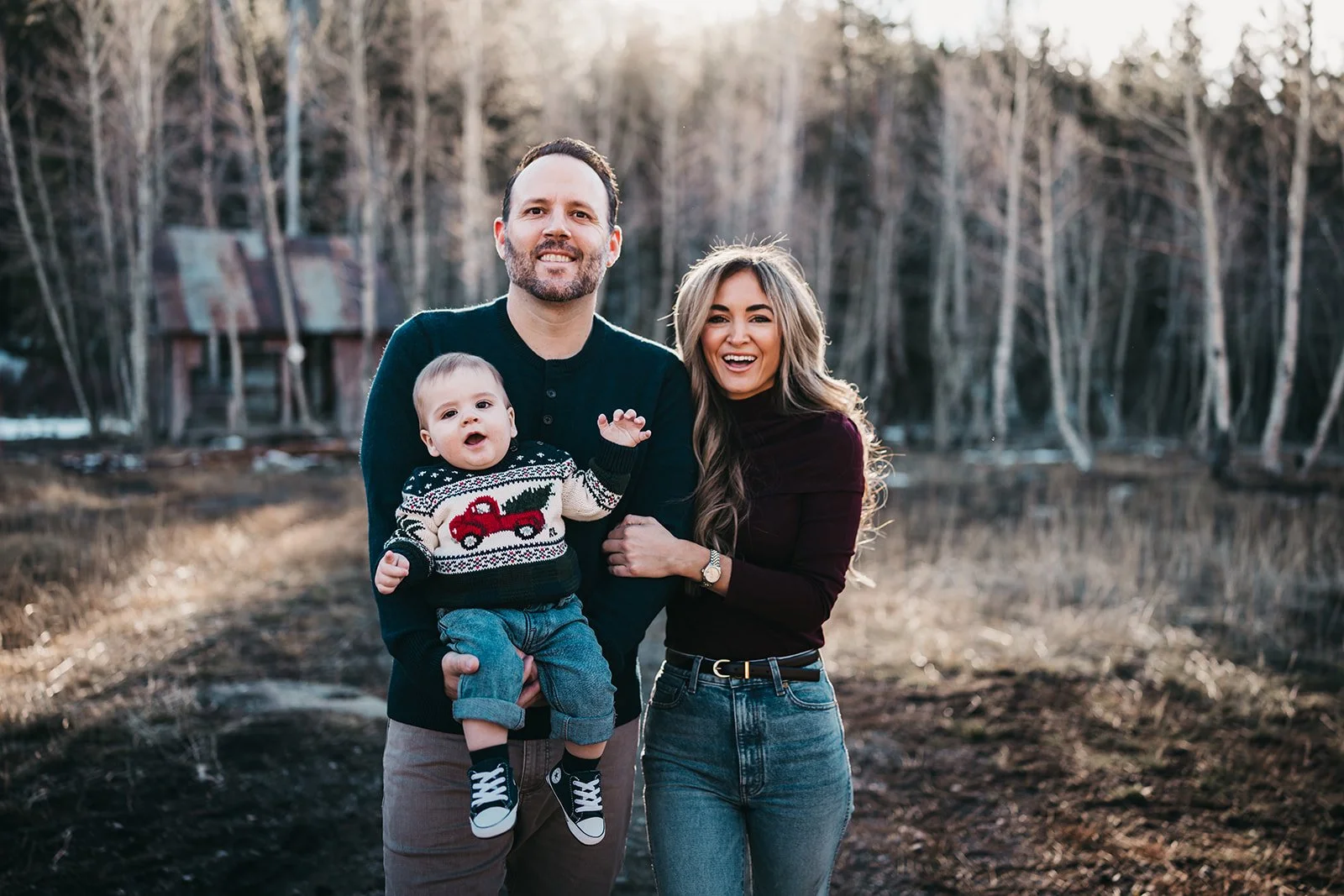 A smiling family of three standing outdoors near a body of water in a wooded area during late afternoon. The man is holding a young boy wearing a sweater with a red fire truck design, and the woman is standing next to them, all appearing happy and en